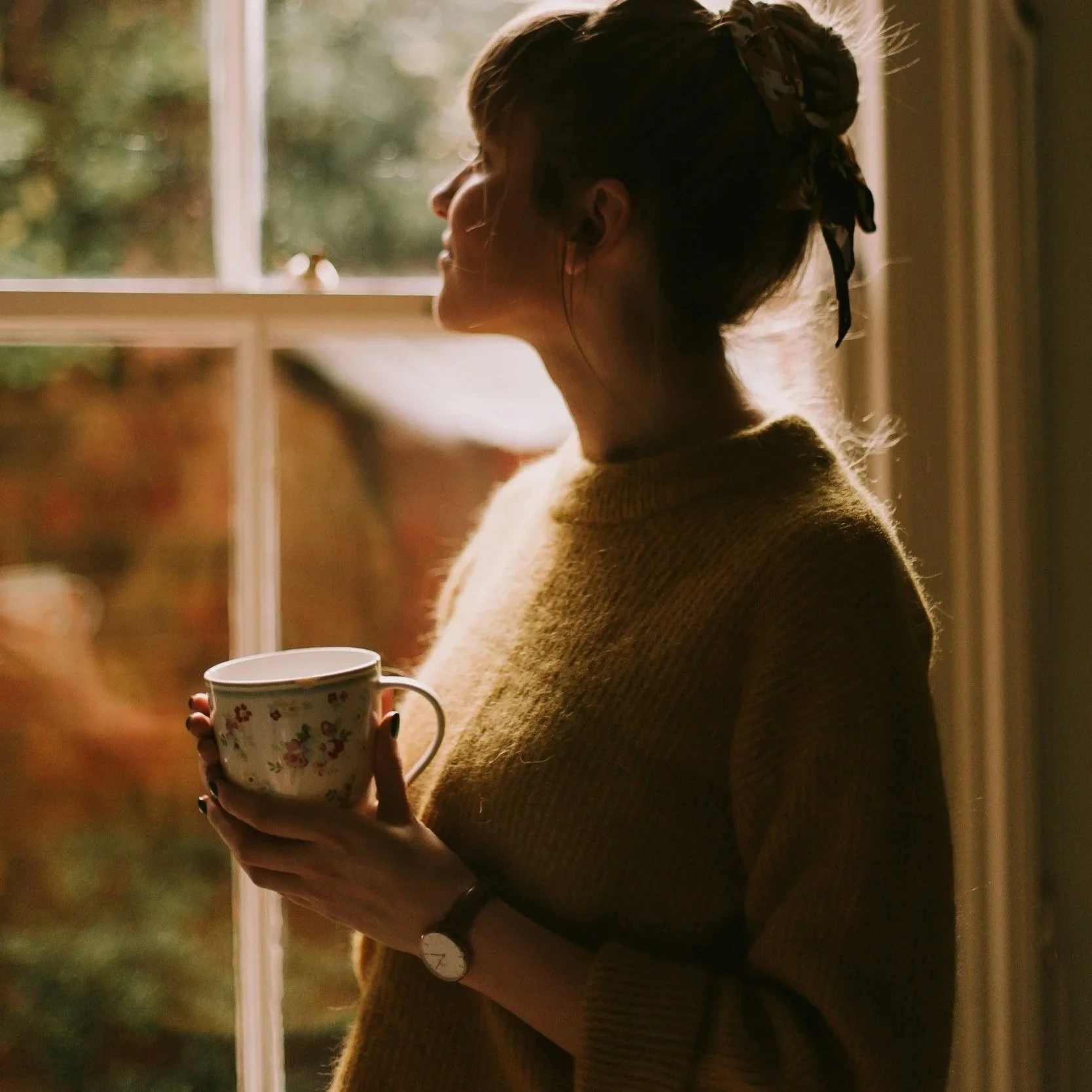A Christian mom standing at the window during the morning, holding a cup of coffee.