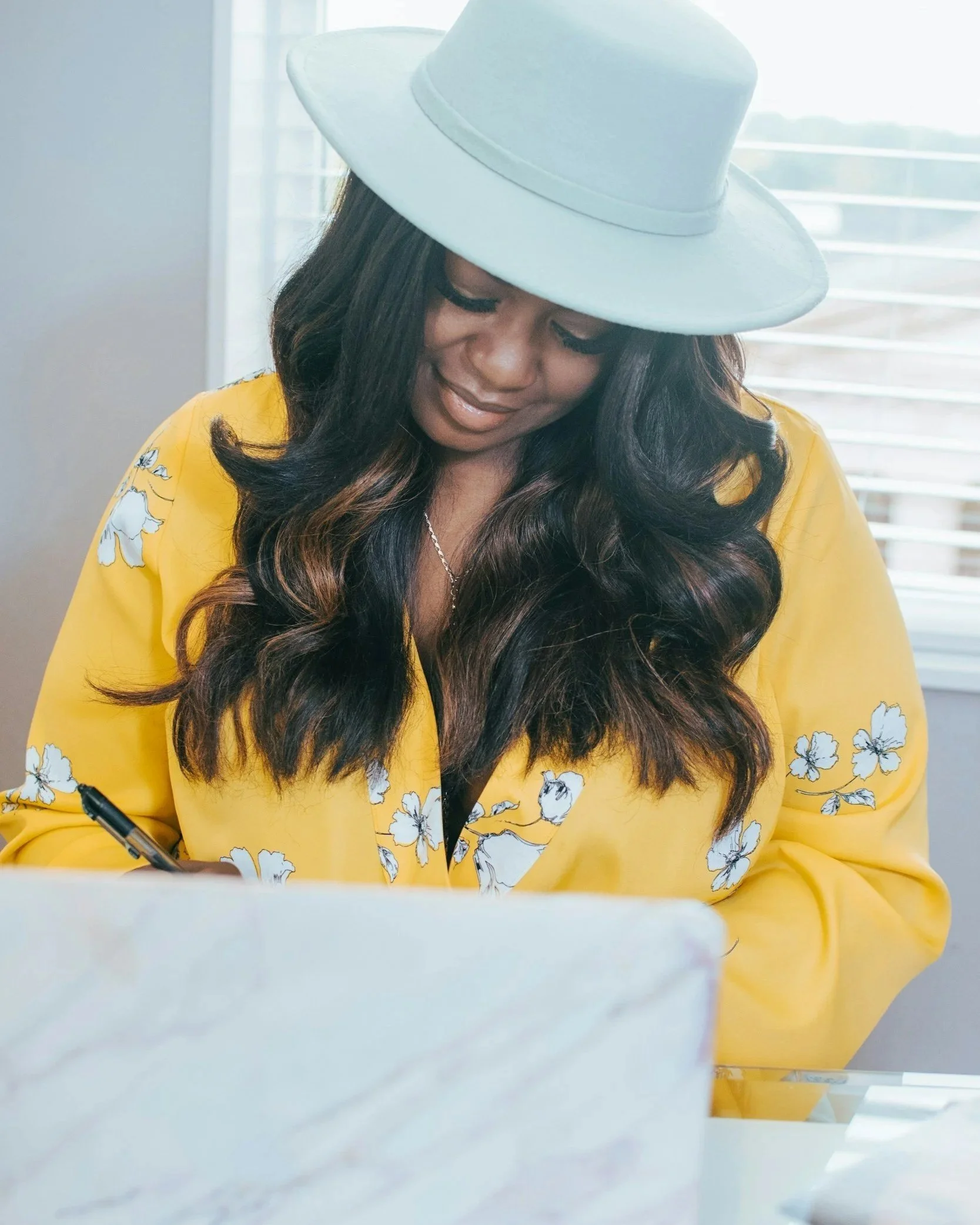 An African American mom sitting at her desk wearing a wide brim hat and yellow blouse.