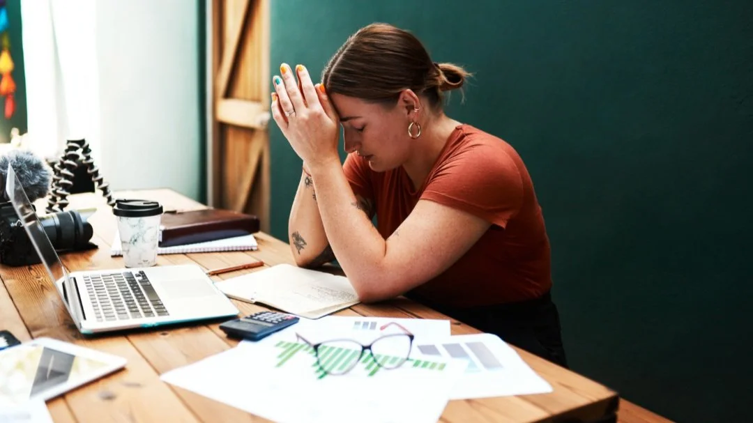 Mom sitting at desk with laptop and coffee. Her hands on on her head and looking down.