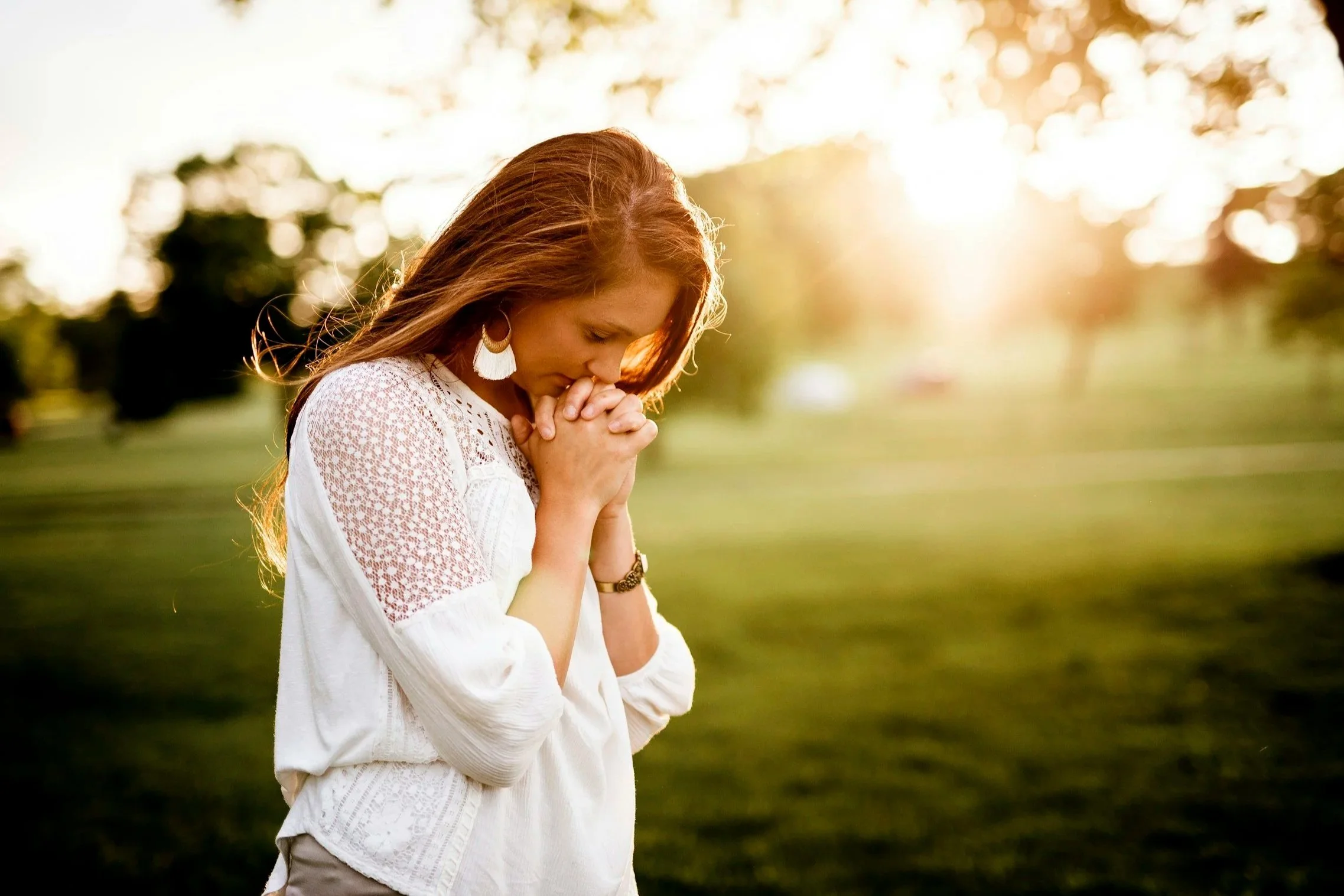 An outside scene with a mom wearing white and standing with her hands in prayer position.