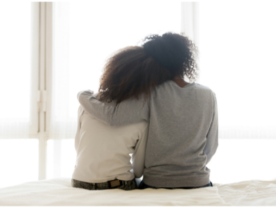 Mom and daughter sitting on edge of bed with moms arm wrapped around the daughter