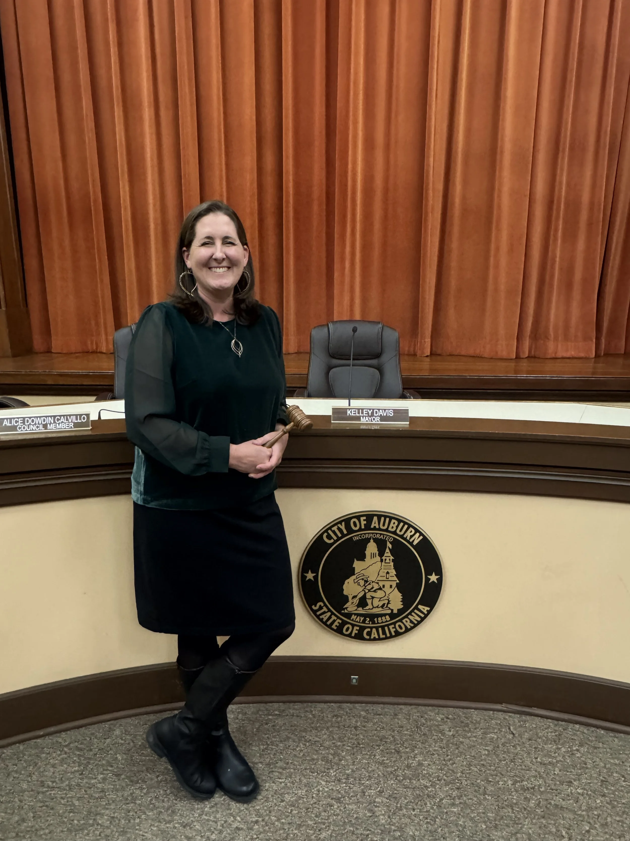A woman standing in front of a city seal at a podium, smiling and holding a gavel, with gold curtains behind her.