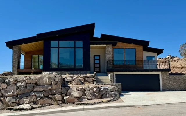 3D rendering of a modern wooden house with large windows, stone accents, and a covered balcony. It features a stone retaining wall and a driveway with a white car parked. The landscaping includes shrubs and trees along the driveway and around the property.