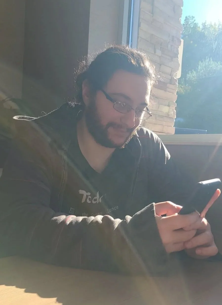 Anthony Ziello sitting at a table, looking at his phone, with sunlight shining in from a window behind him.