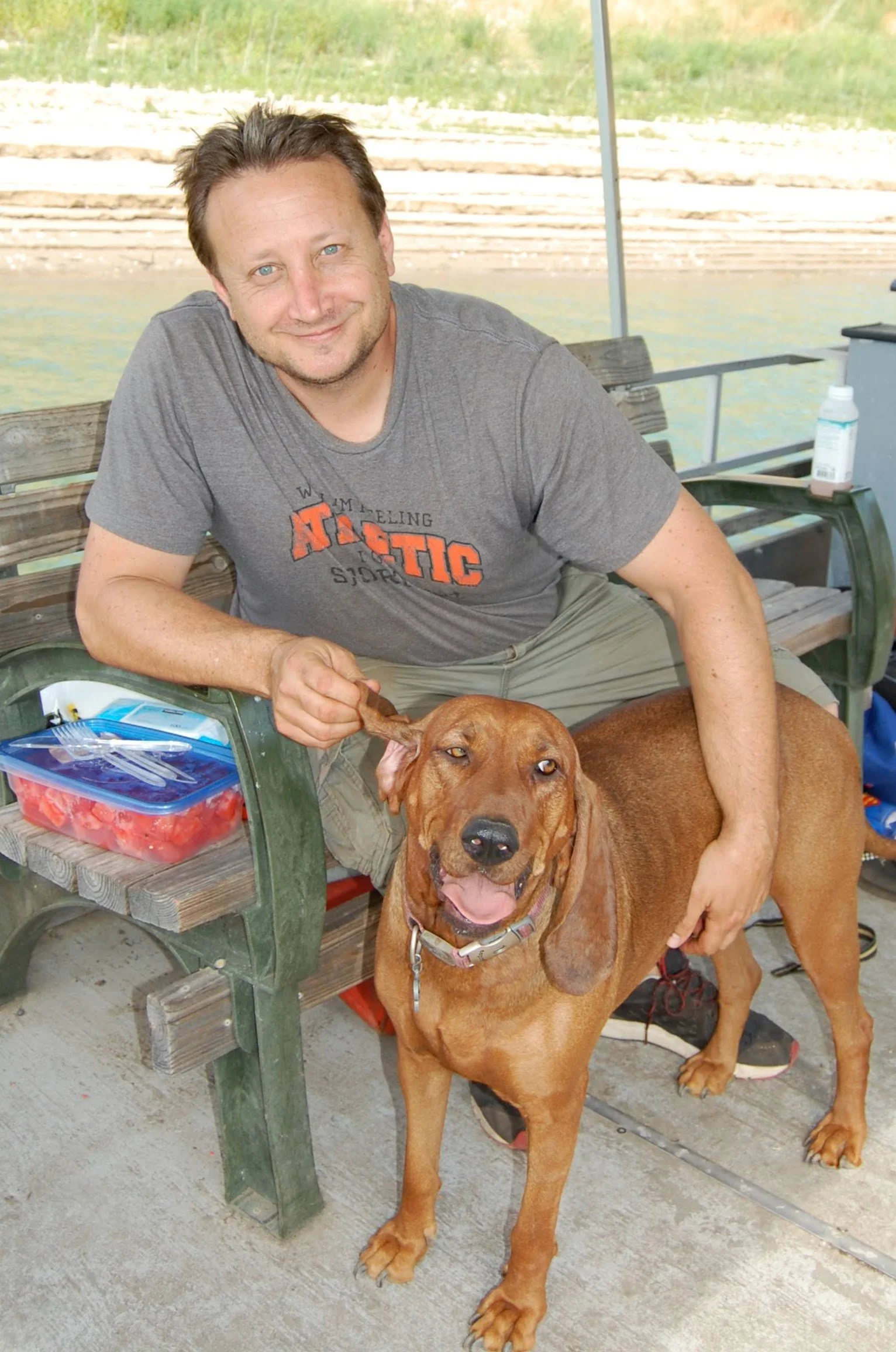 Man sitting on bench with hands on redbone coonhound dog.