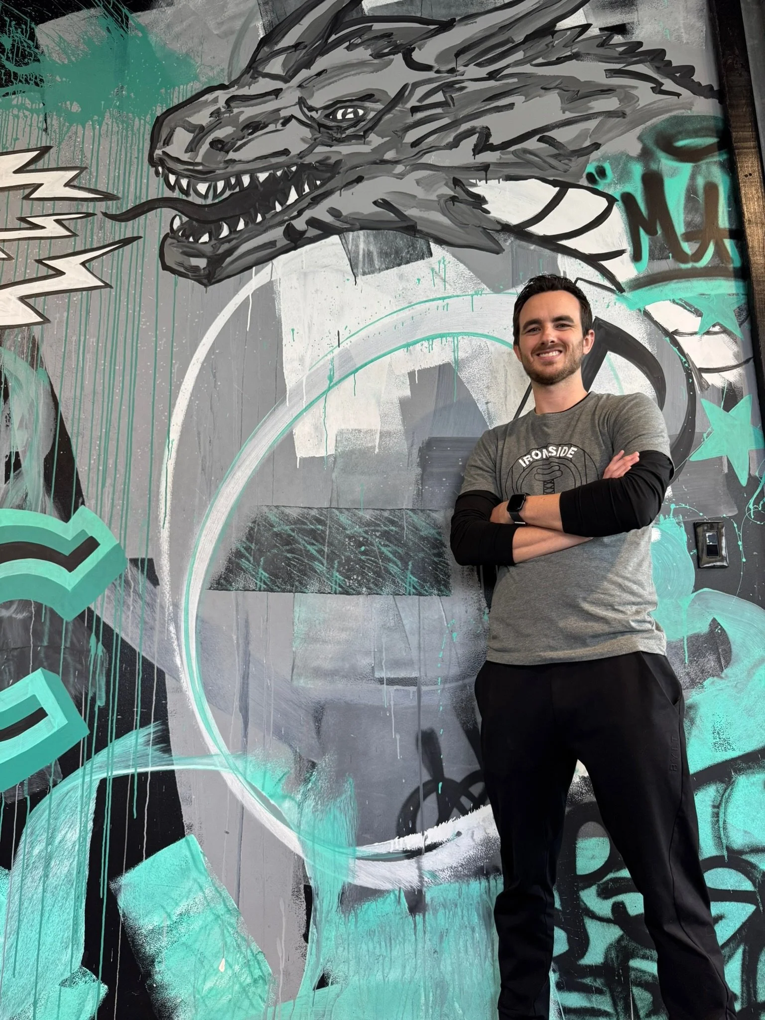 A man with arms crossed, smiling, standing in front of a colorful graffiti mural featuring a dragon's head, stars, and abstract designs.