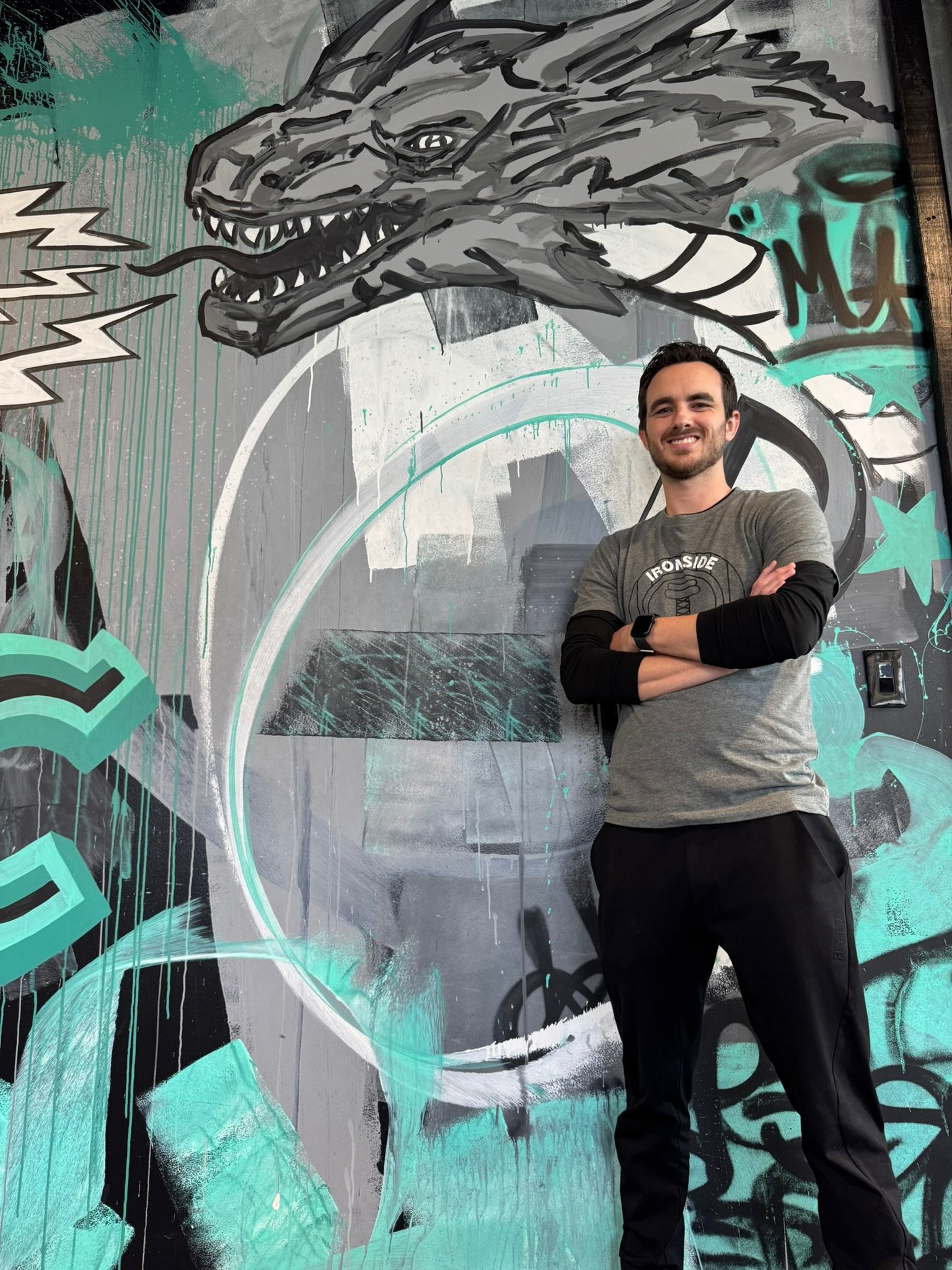 A smiling man with crossed arms standing in front of a colorful graffiti mural featuring a dragon head, geometric shapes, and abstract designs in shades of teal, black, white, and gray.