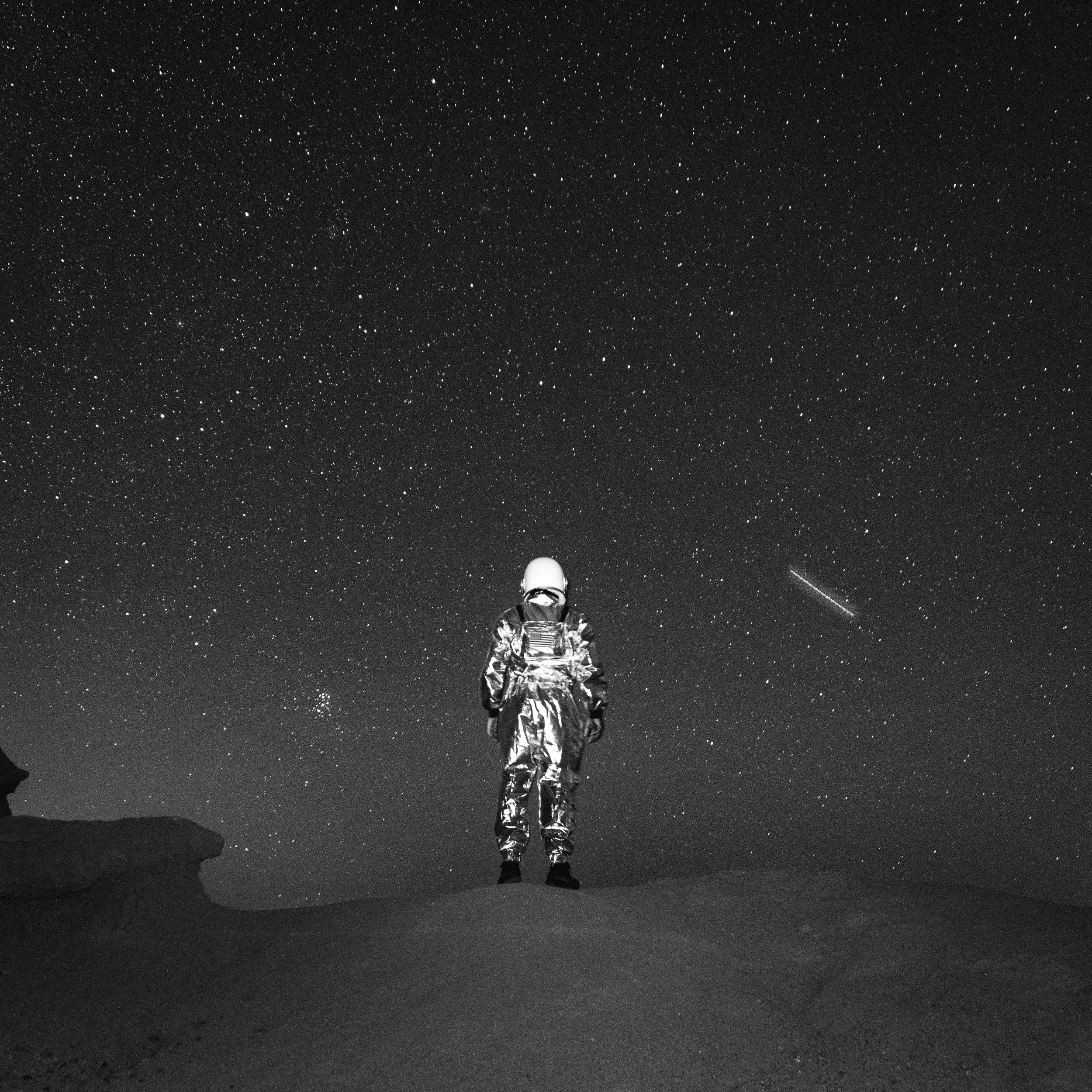 Astronaut standing on a hill in a space suit, looking up at a starry night sky with a visible galaxy and shooting star.