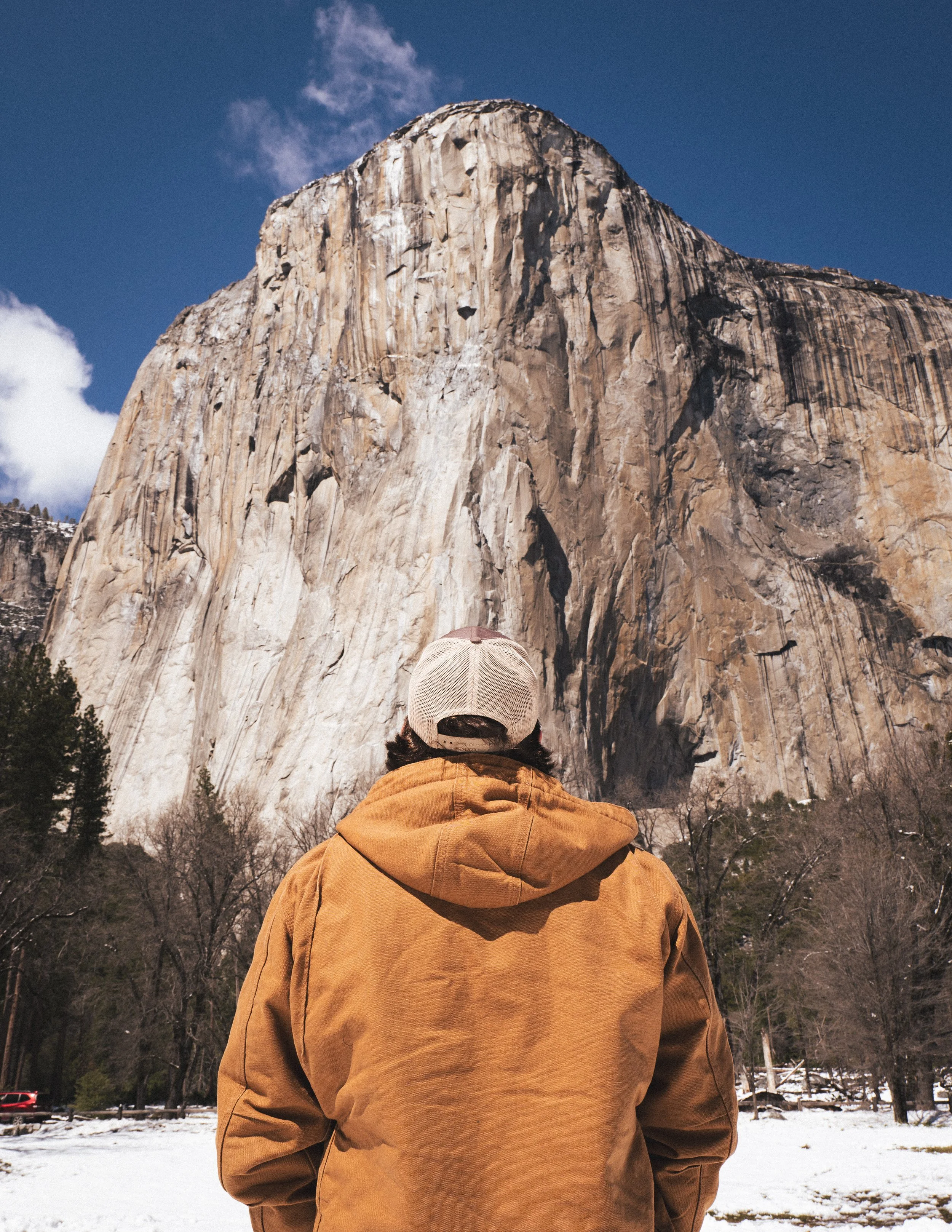 A person in a brown jacket and beige cap standing outdoors in front of El Capitan mountain in Yosemite National Park.