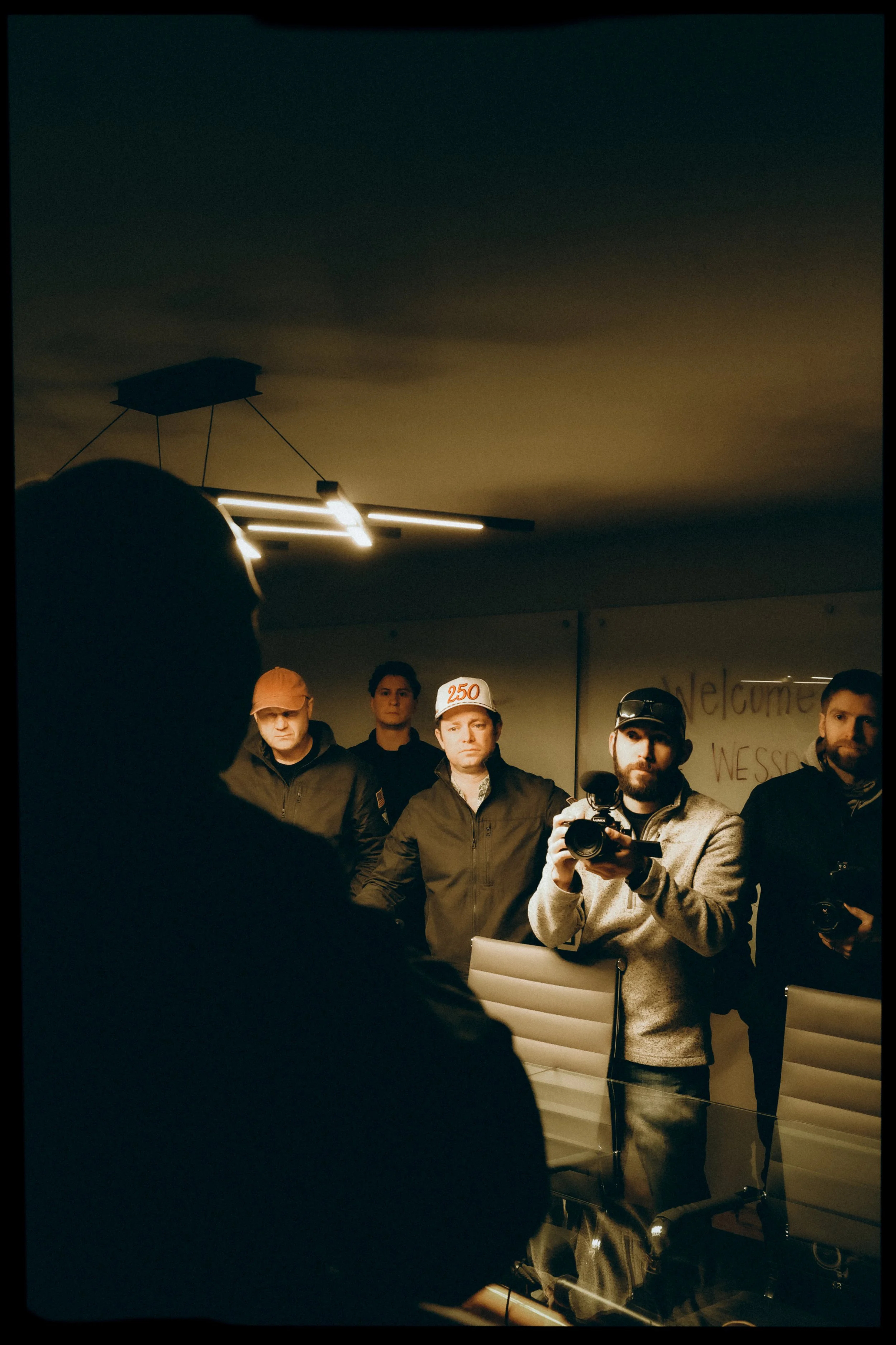 Group of five men standing indoors, one man in the foreground holding a camera, wearing a grey hoodie and a cap, with others standing behind him, some wearing jackets and glasses.