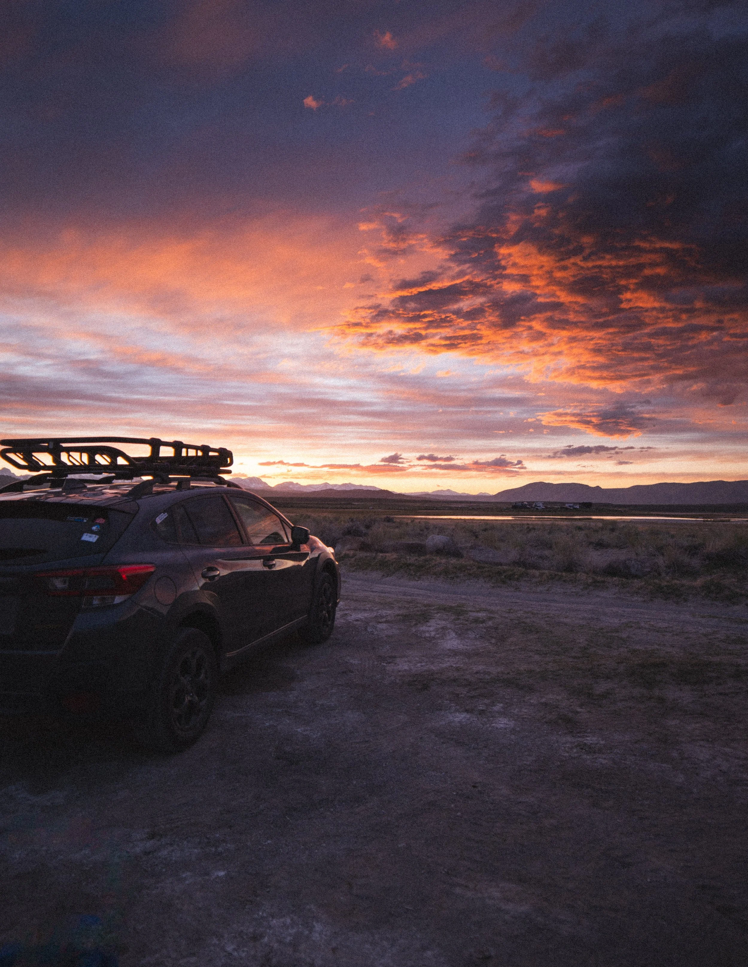 A black SUV with a roof rack parked on a dirt road at sunset with a colorful sky and mountains in the distance.