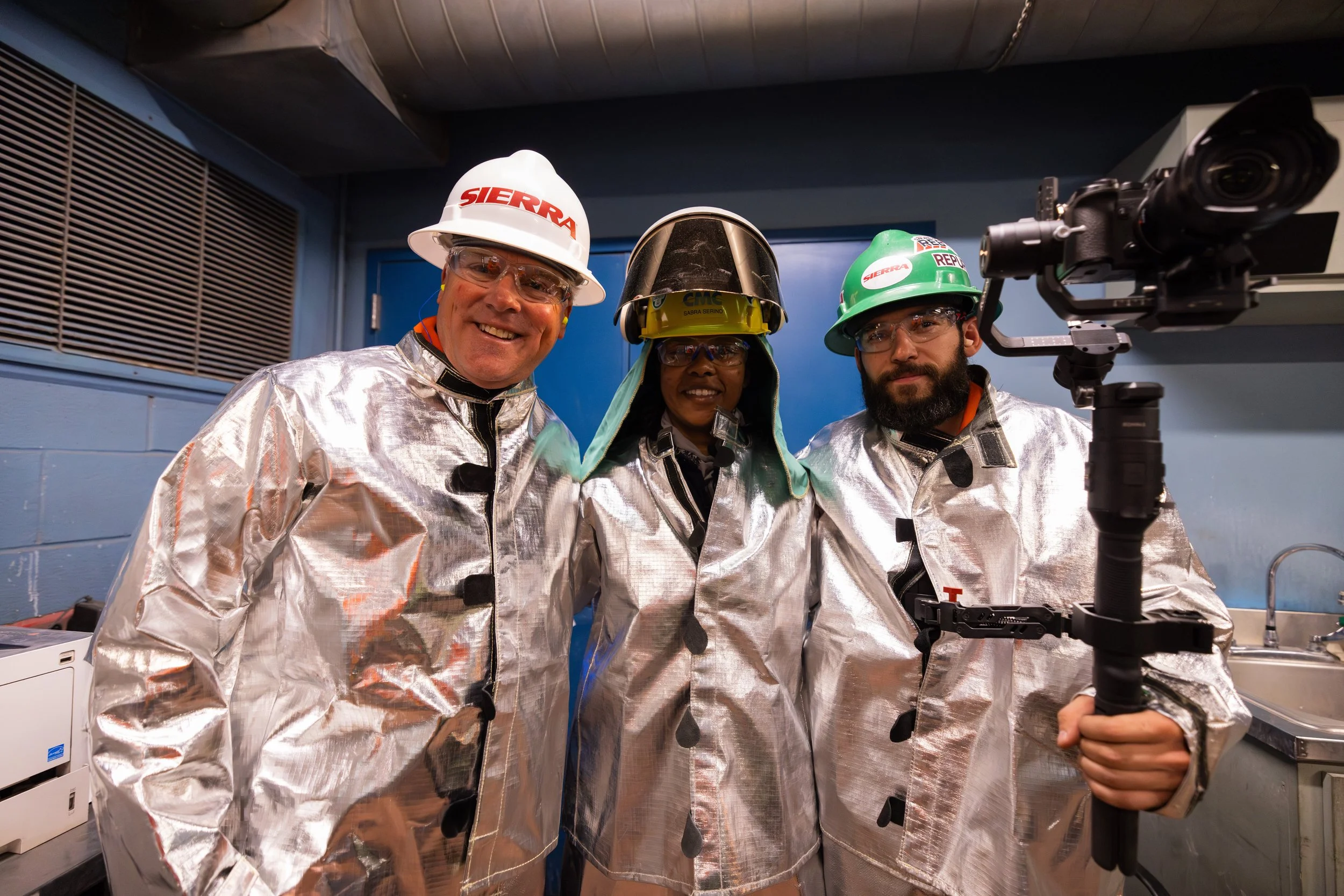 Three workers wearing safety helmets, protective glasses, and shiny metallic suits posing together in an industrial room. One is holding a camera mounted on a stabilizer.