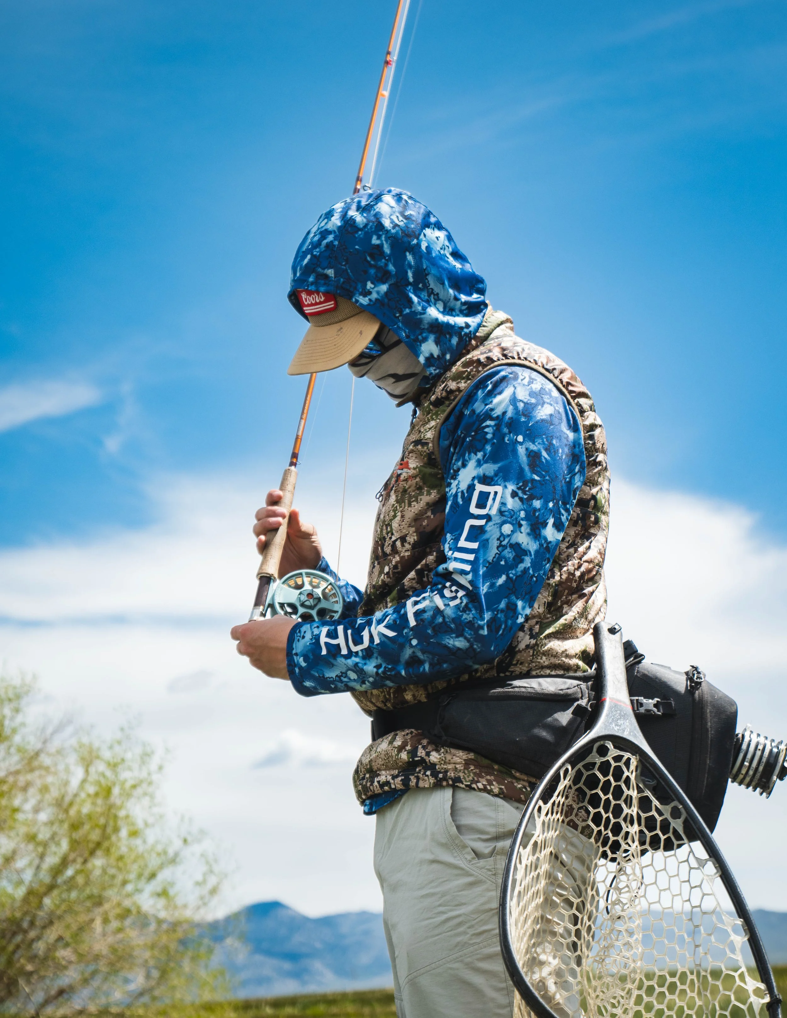 Person in camouflage and blue hoodie fishing outdoors with a fly rod and net under a partly cloudy sky.