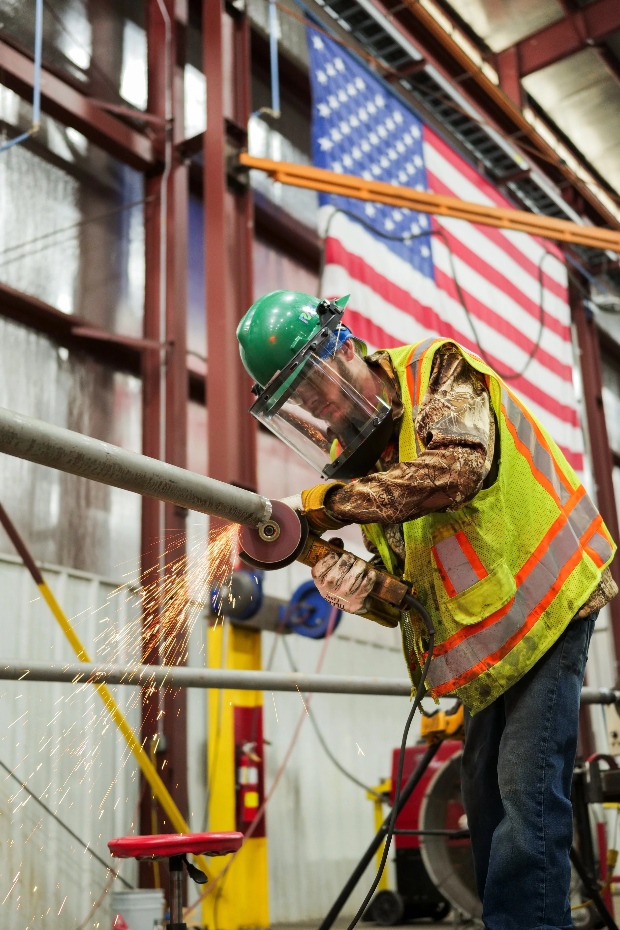 A worker in safety gear, including a green helmet and face shield, is grinding a metal pipe, producing sparks, inside an industrial facility with American flags hanging in the background.