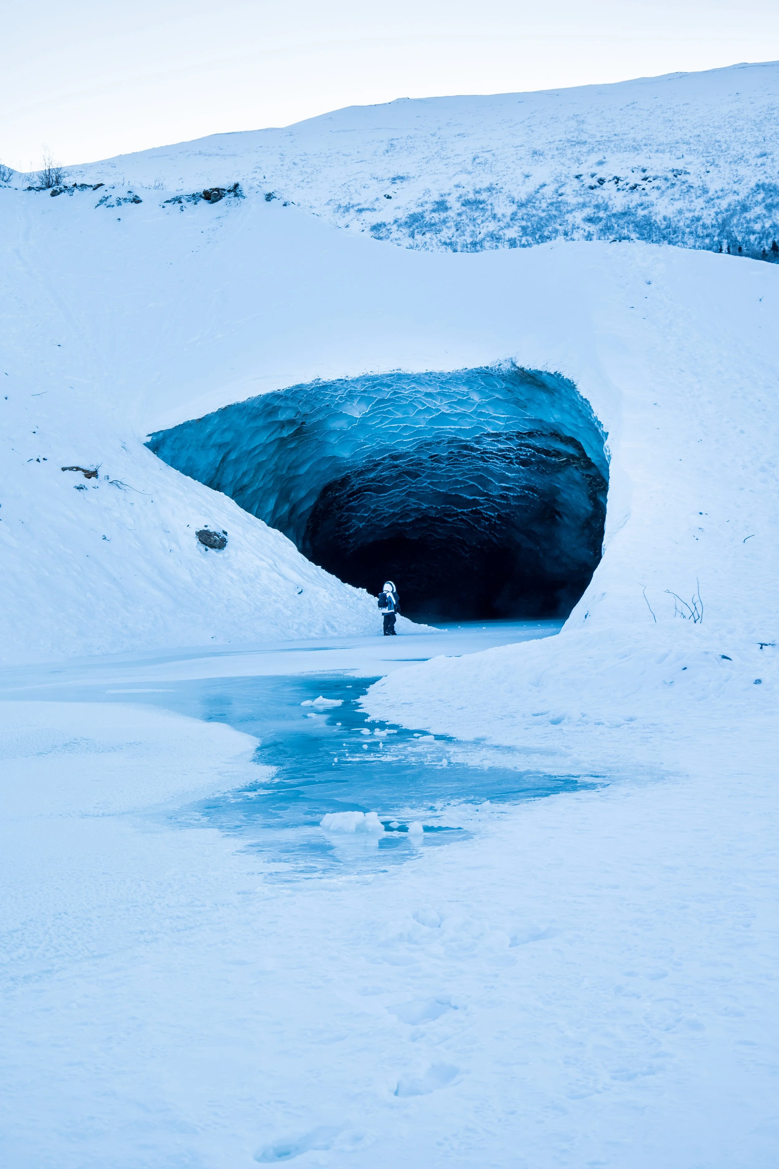 A person standing in front of a large ice cave in a snowy landscape with mountains in the background.