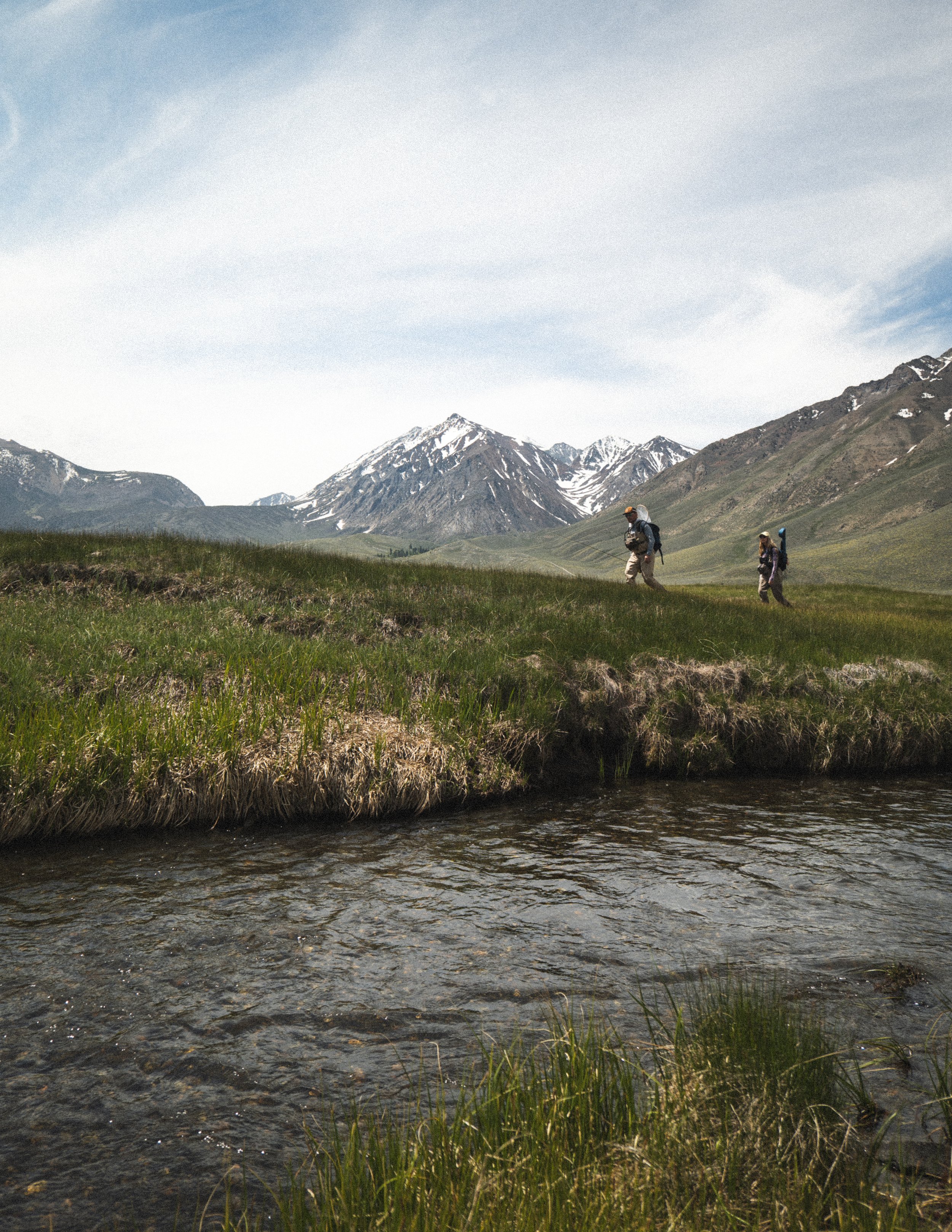 Two hikers walking along a grassy trail above a small creek with snow-capped mountains in the background.