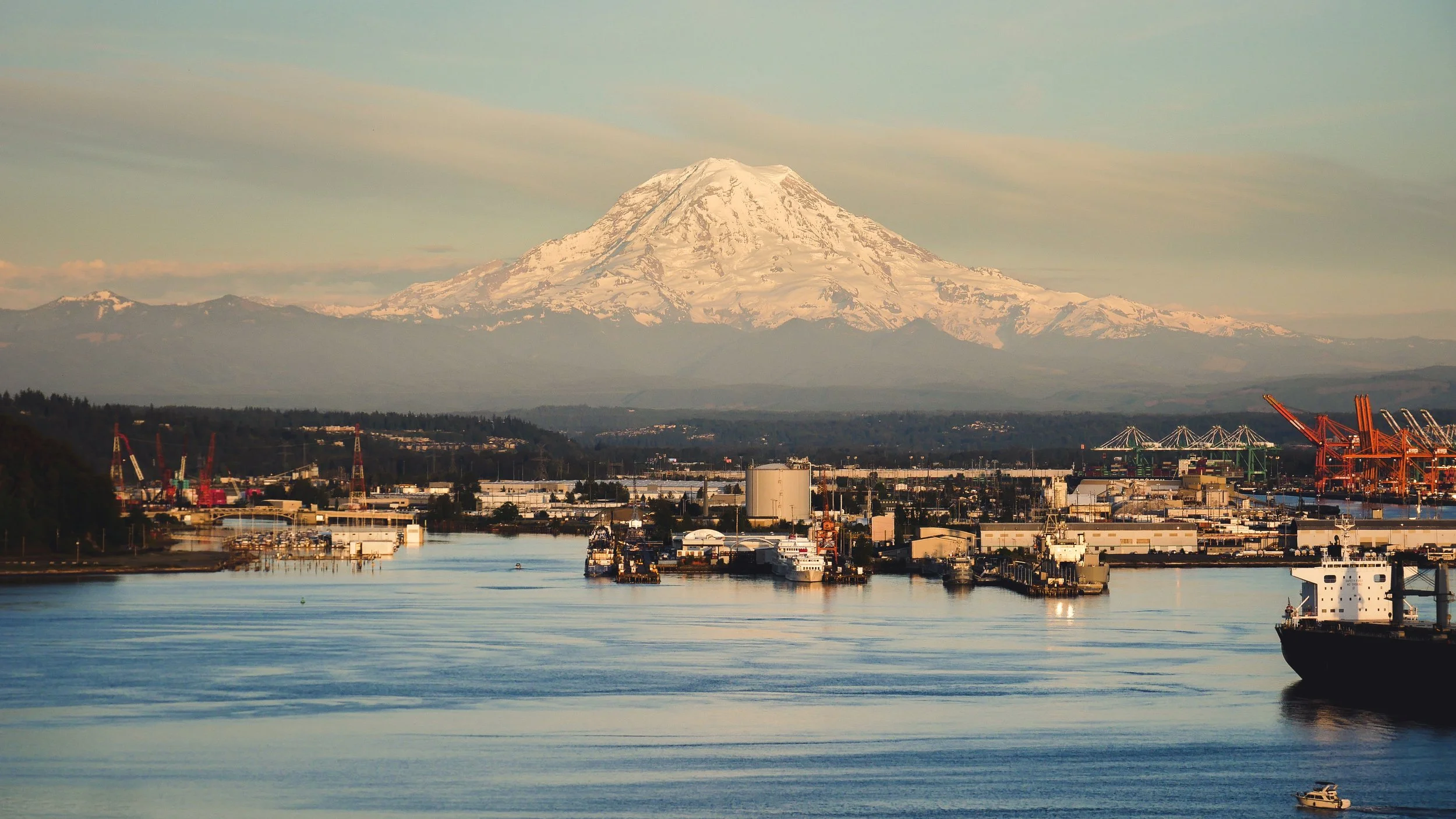 Snow-capped Mount Rainier in the background over an industrial harbor scene with cranes, warehouses, boats, and water in the foreground.