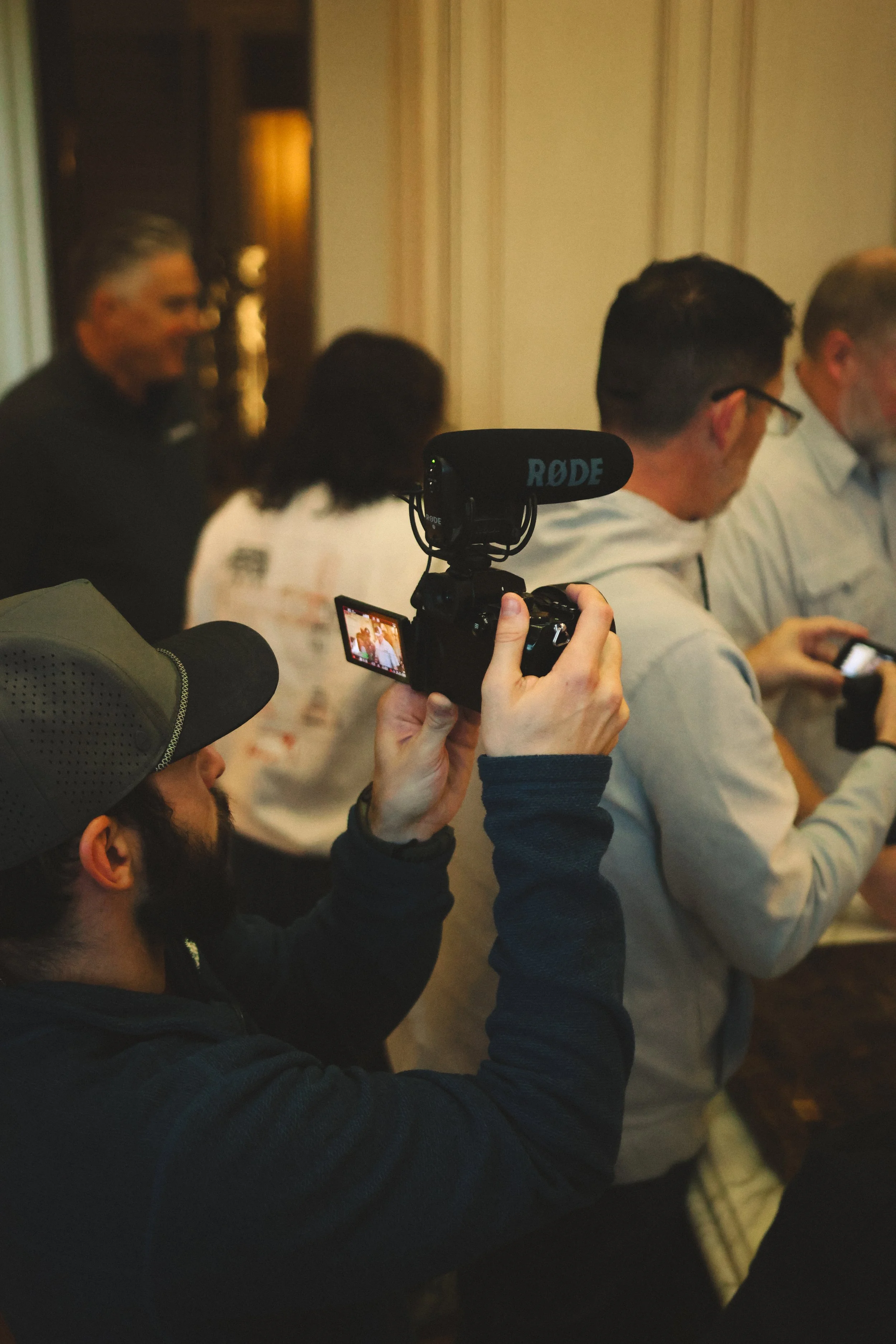 Person (Andrew Sacchini) taking a photo with a camera with a Rode microphone in a room with several people standing and talking.