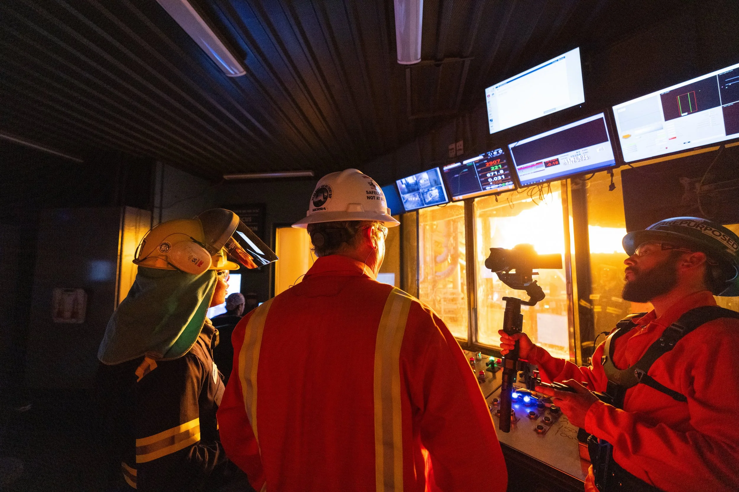 Industrial workers monitoring a large plant in a control room with multiple screens showing data and security footage.