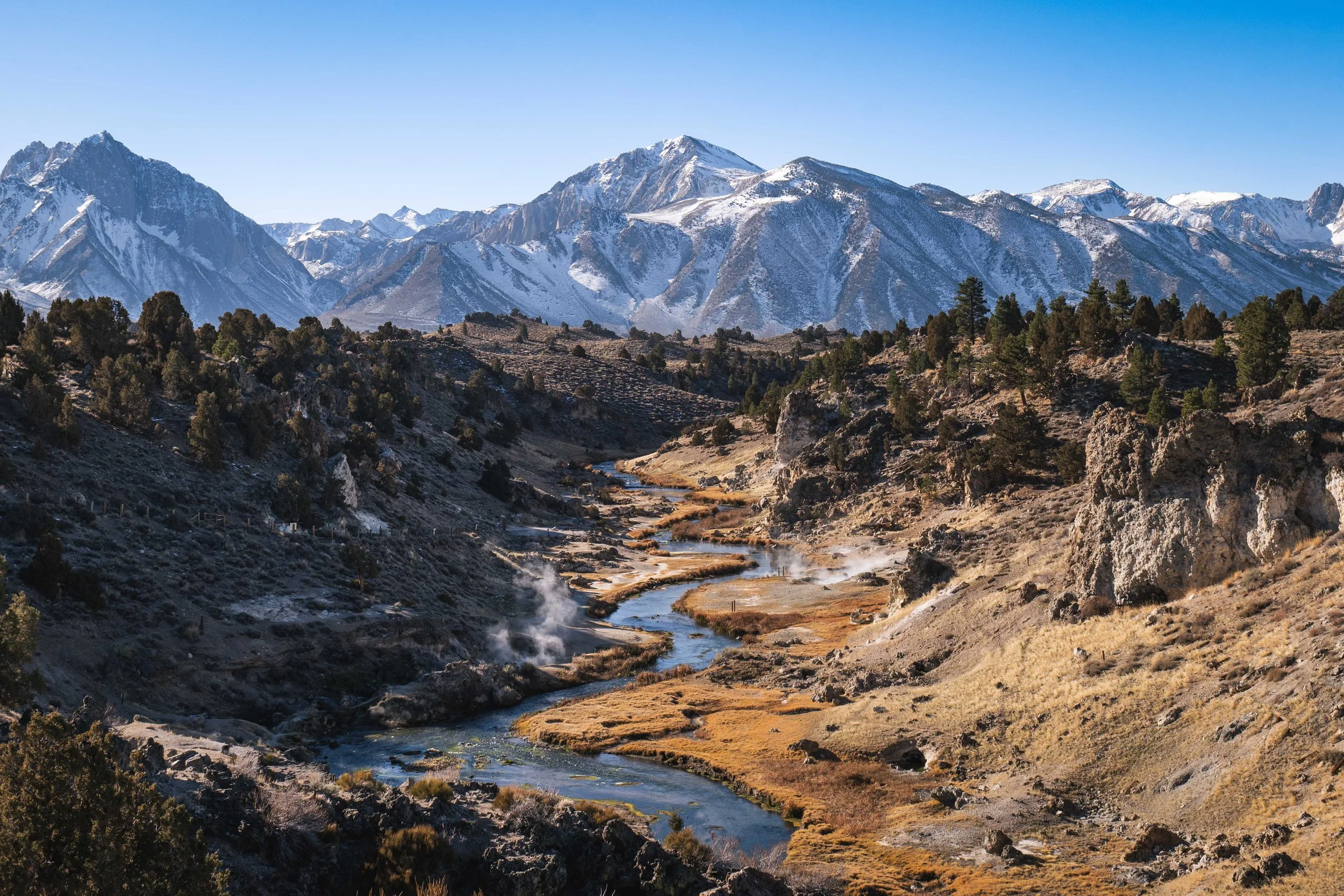 Snow-capped mountains behind a valley with a winding river, scattered trees, and steam rising from hot springs