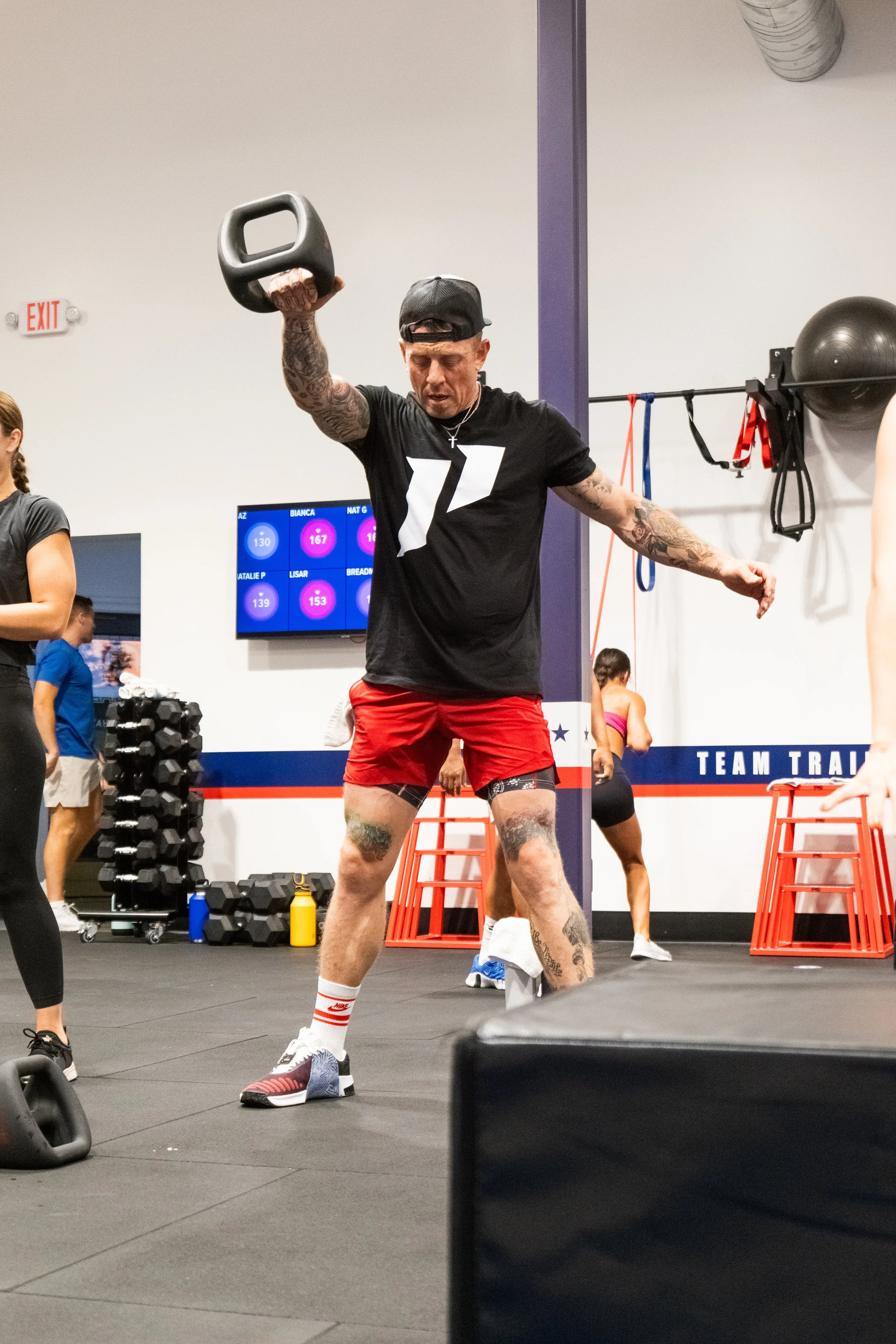 A man in a black T-shirt, red shorts, and a black cap is lifting a kettlebell in a gym, surrounded by other people exercising and fitness equipment.