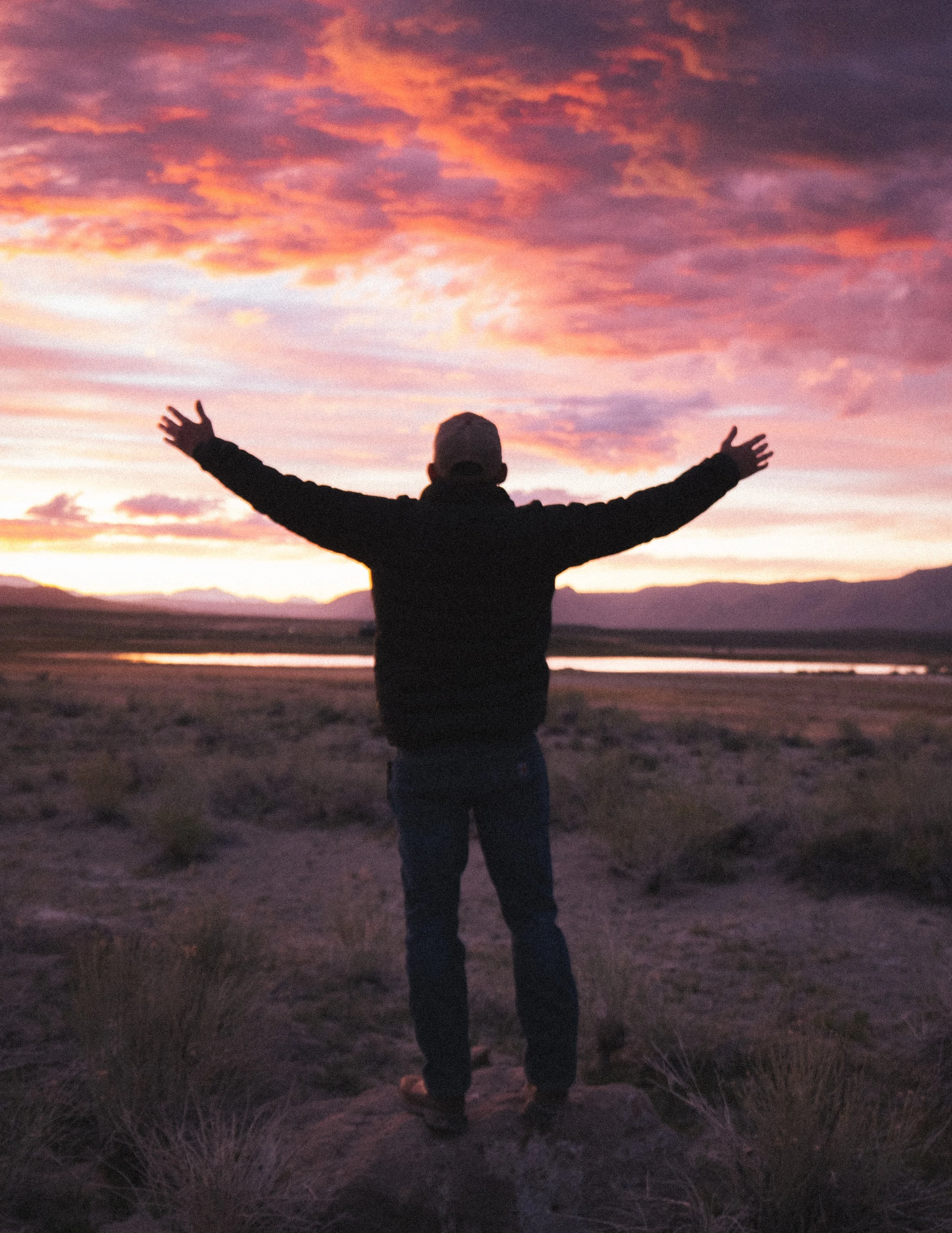 A person standing outdoors at sunset with arms outstretched, overlooking a landscape with mountains and a river.