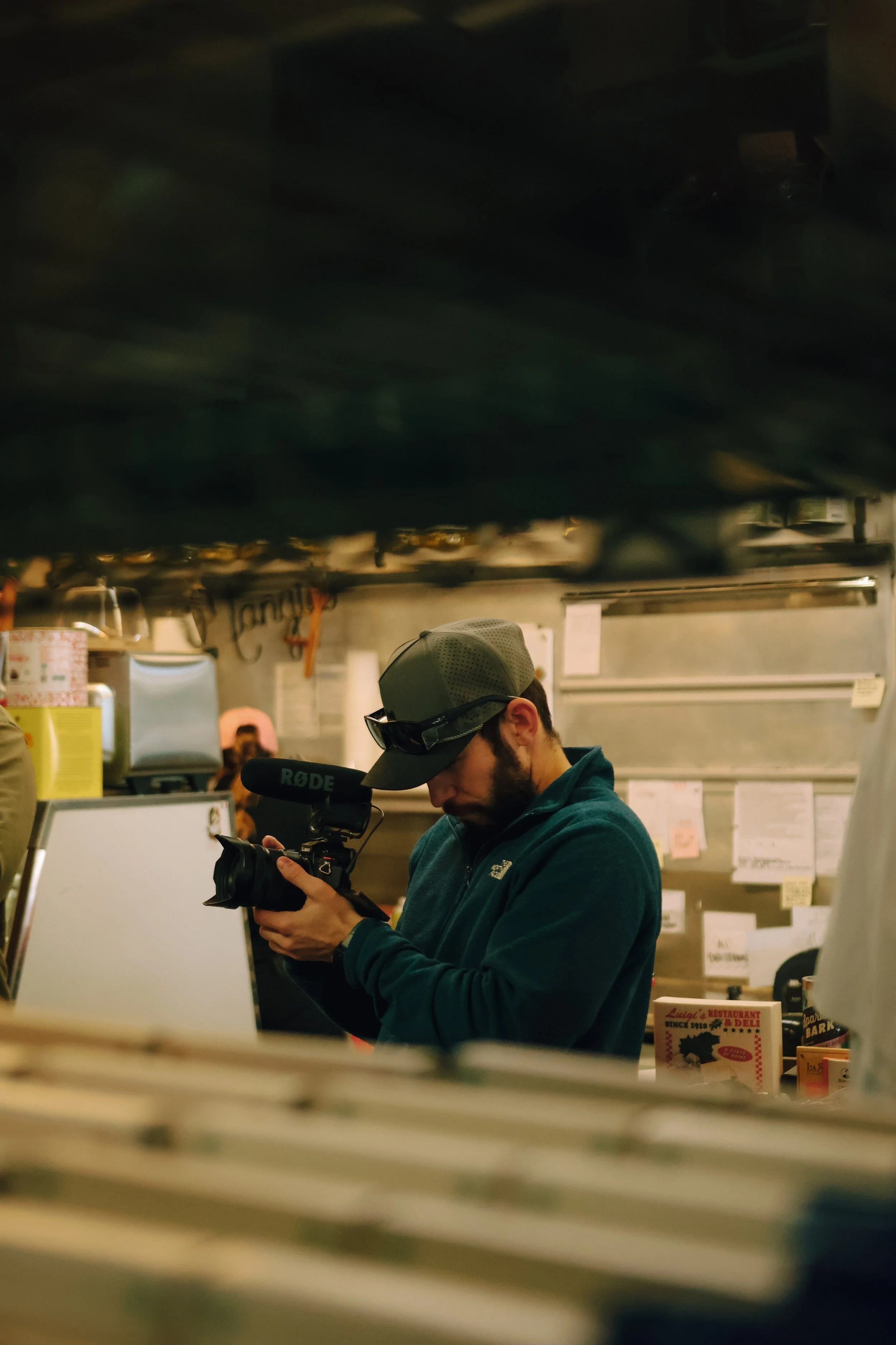 A man (Andrew Sacchini) wearing a cap and glasses, holding a camera with a microphone attached, standing inside a restaurant or deli, with blurry signs and papers on the wall behind him.