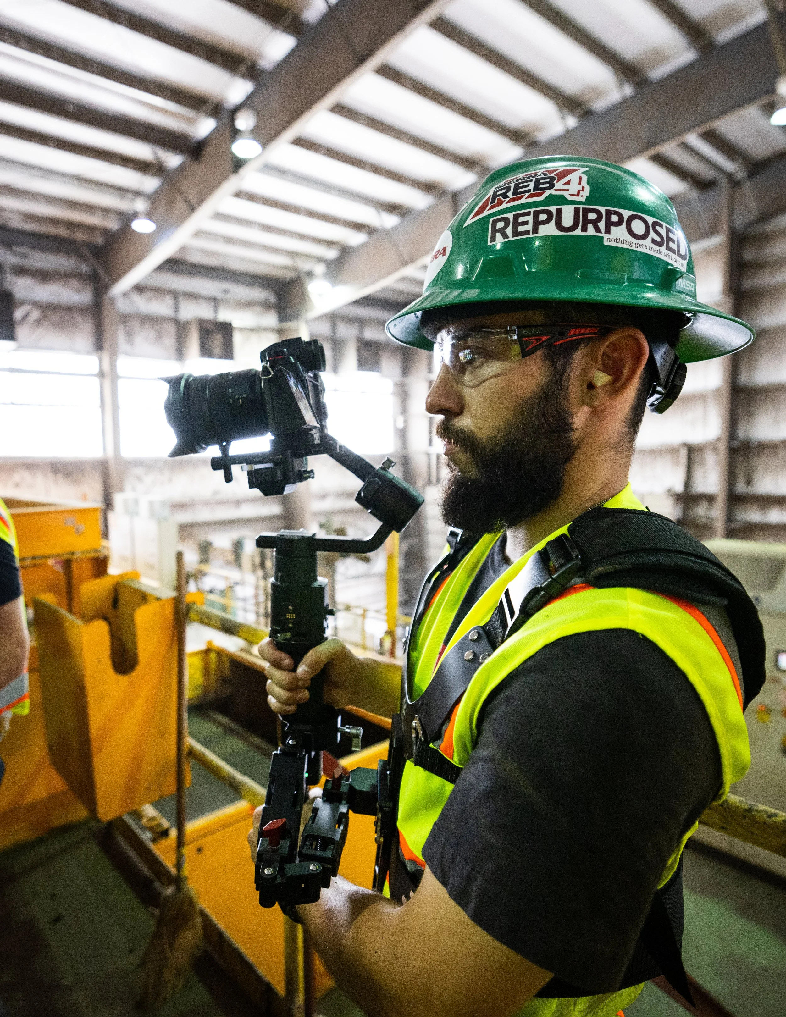 A man (Andrew Sacchini) wearing safety glasses, a green hard hat with 'REBUILD REB4' and 'REPURPOSE' stickers, and a high-visibility vest, holding a camera stabilizer inside an industrial building.