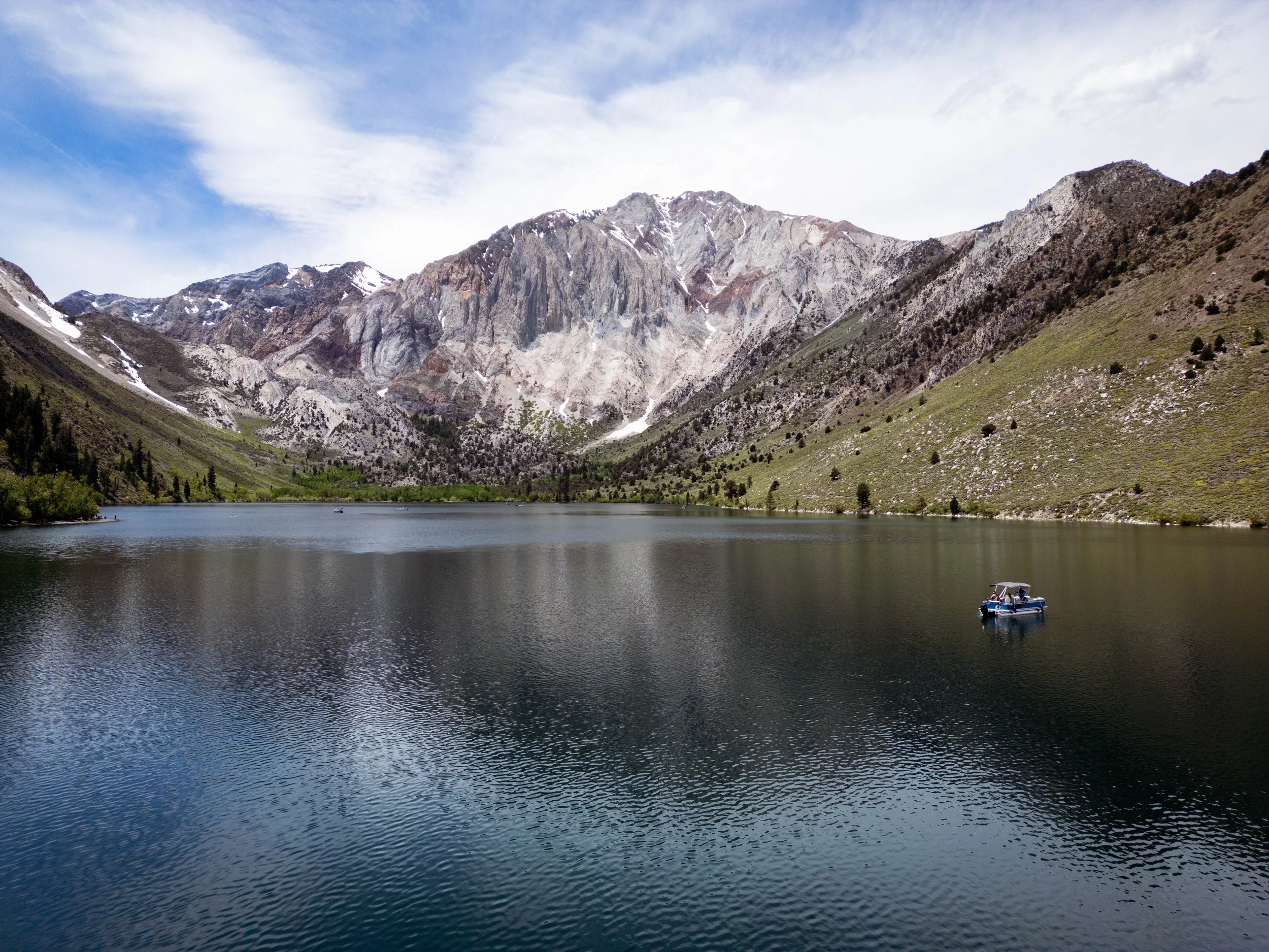 A scenic mountain lake surrounded by tall, rocky mountains and green slopes, with a small boat floating on the calm water under a partly cloudy sky.