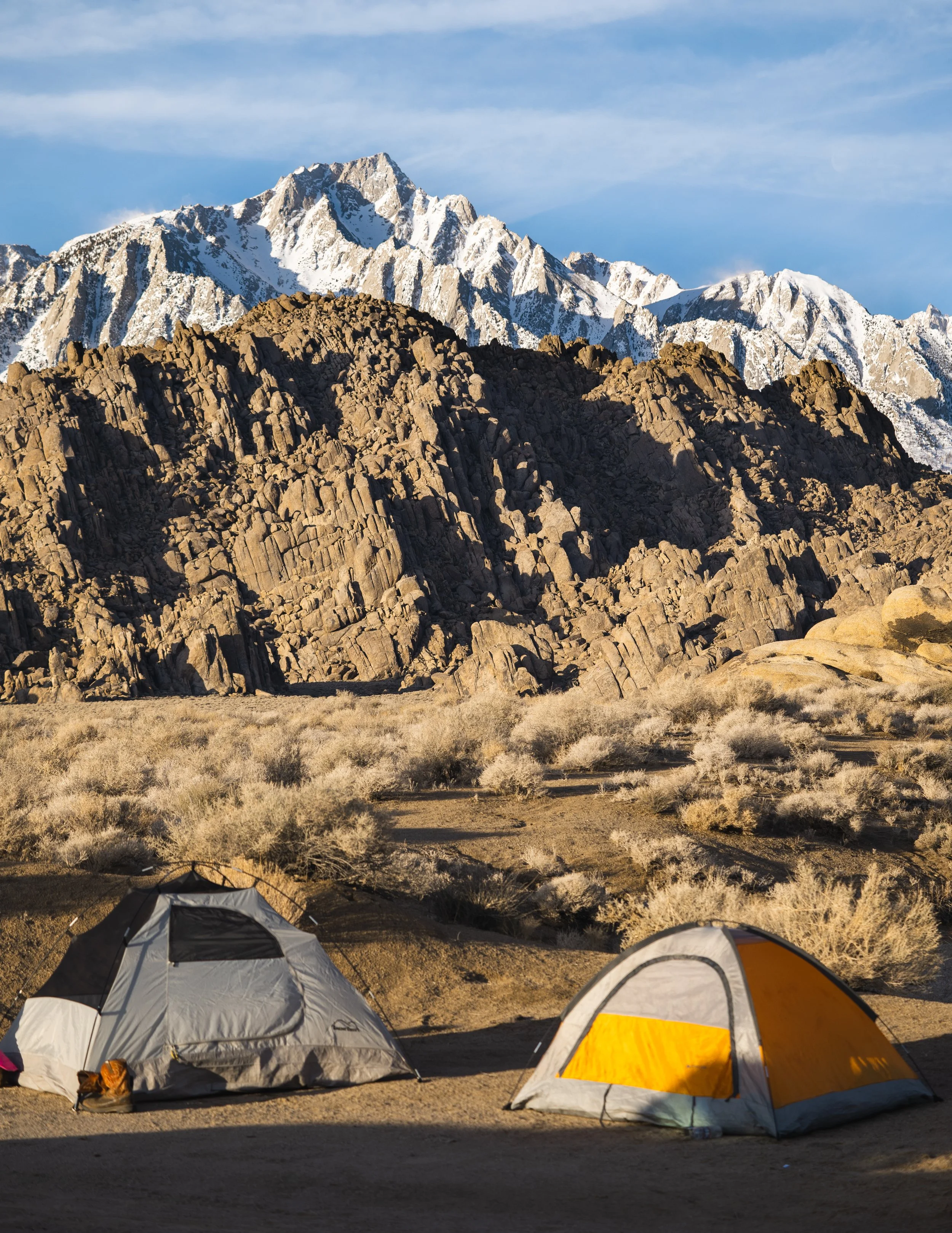 Two tents set up in a desert landscape with rocky mountains and snow-capped peaks in the background.