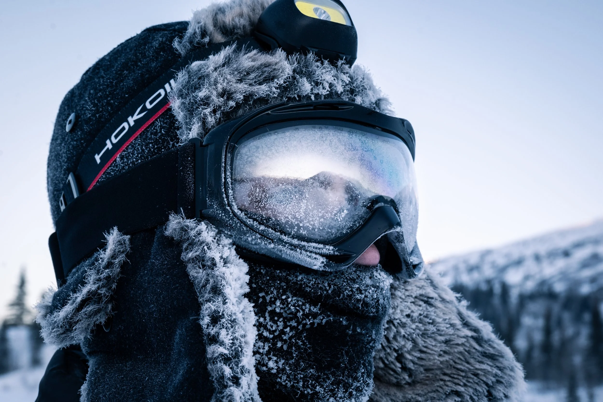 Close-up of a person wearing a ski helmet, goggles, and a fur-lined winter coat in a snowy outdoor setting.