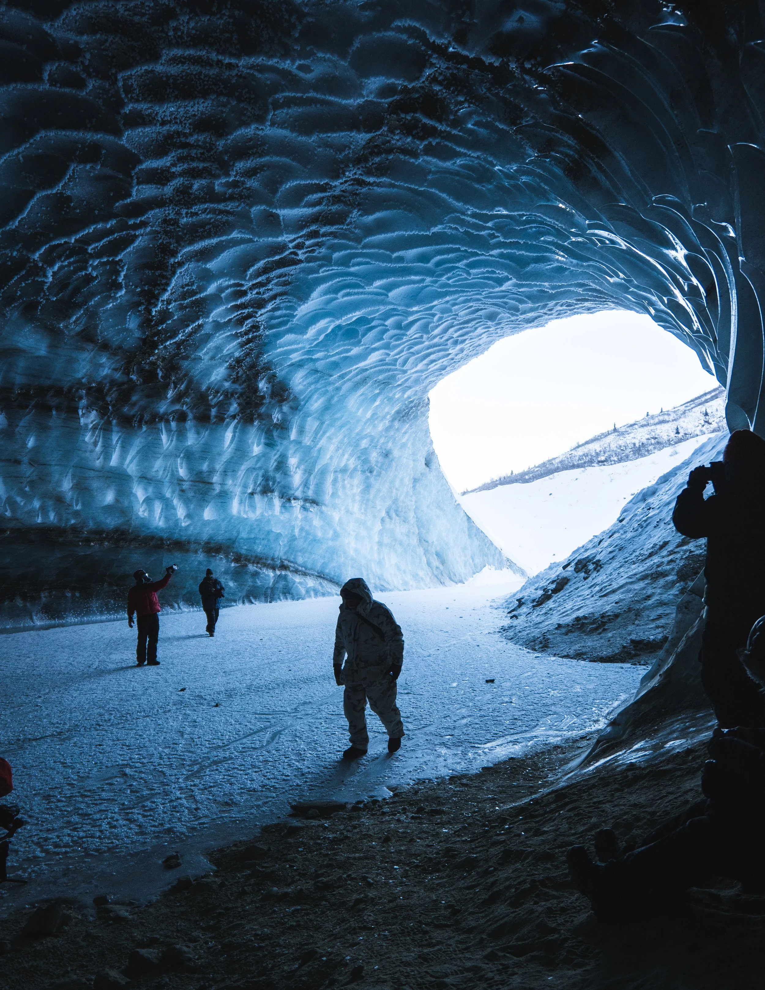 People exploring an icy ice cave with blue crystal formations on the ceiling and snow-covered landscape outside the cave entrance.