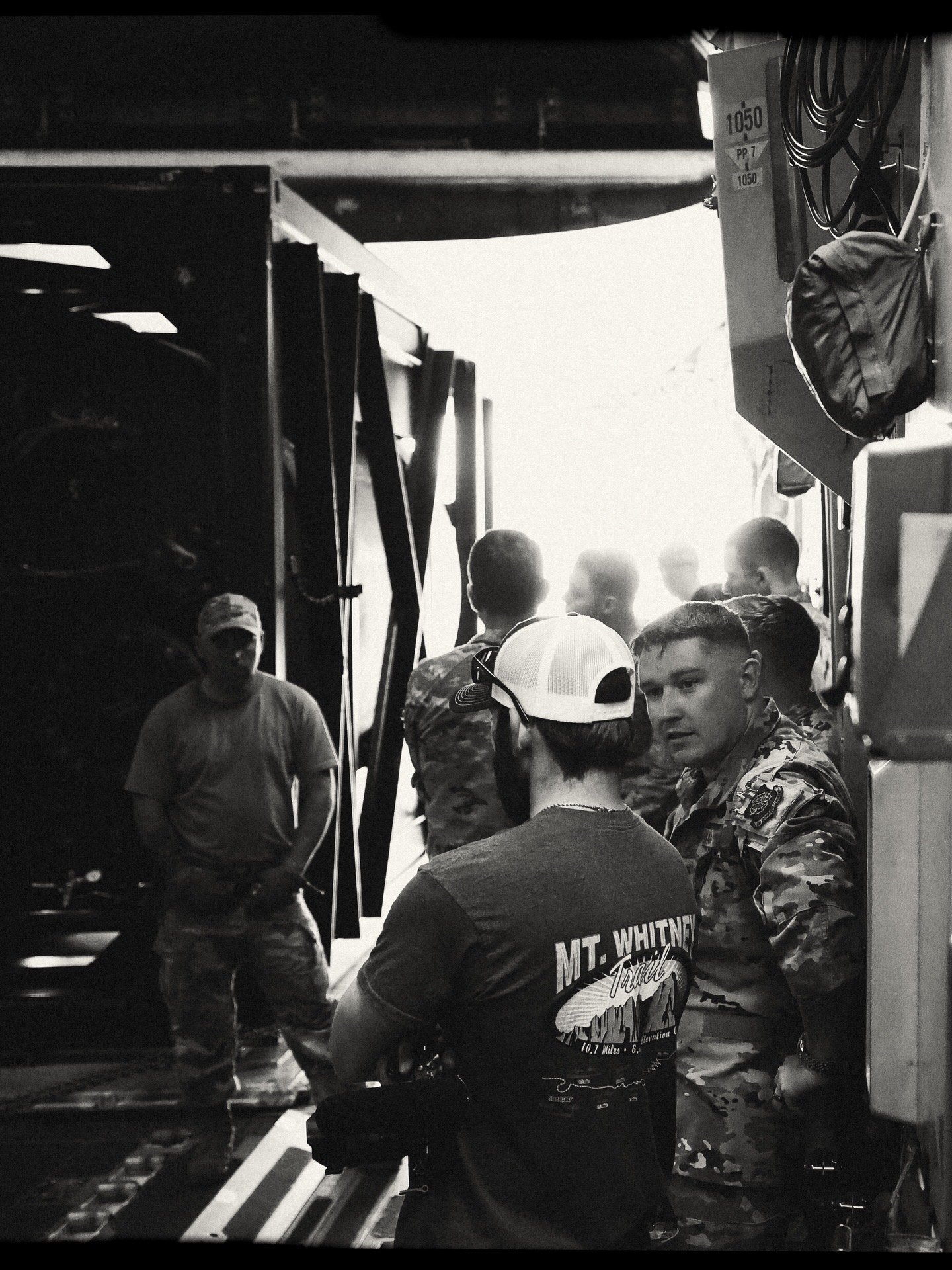 Group of soldiers in military uniform inside a loading area, standing near equipment, with bright light in the background.