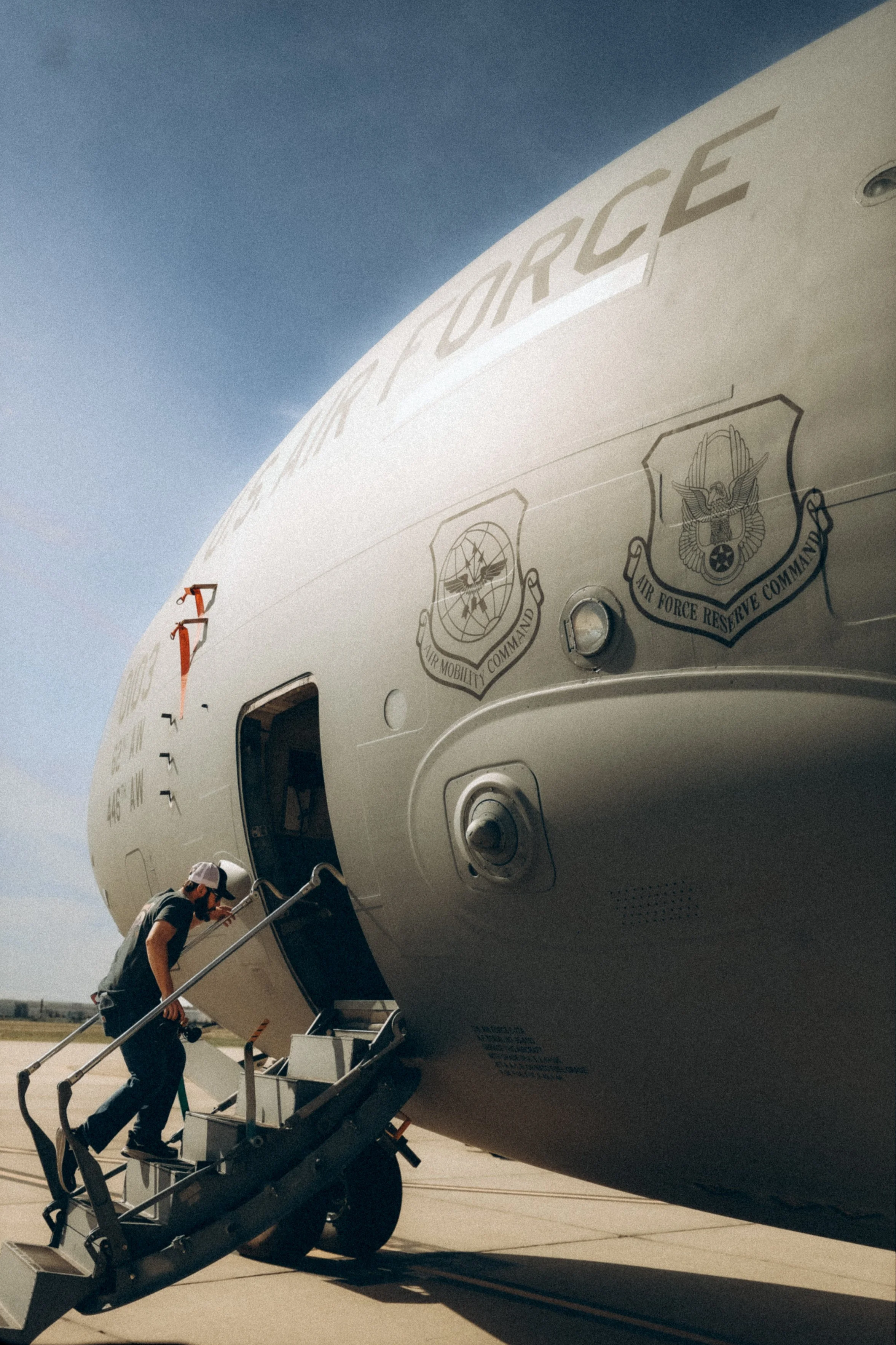 A person (Andrew Sacchini) boarding a U.S. Air Force aircraft via a mobile staircase, with insignias and text visible on the aircraft fuselage.