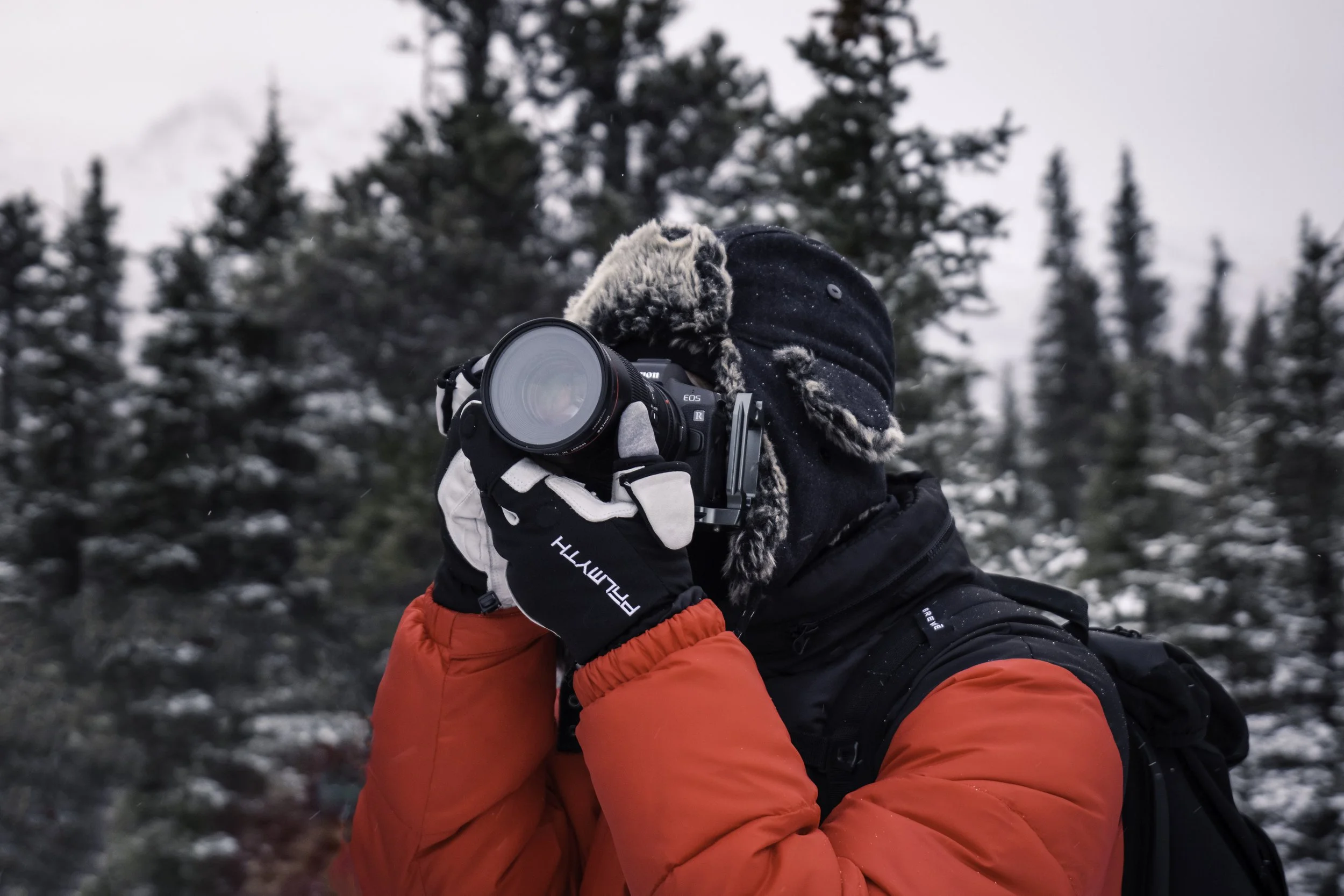 Person dressed in a winter coat and gloves, holding a camera with a large lens, taking a photograph outdoors in a snowy forest.