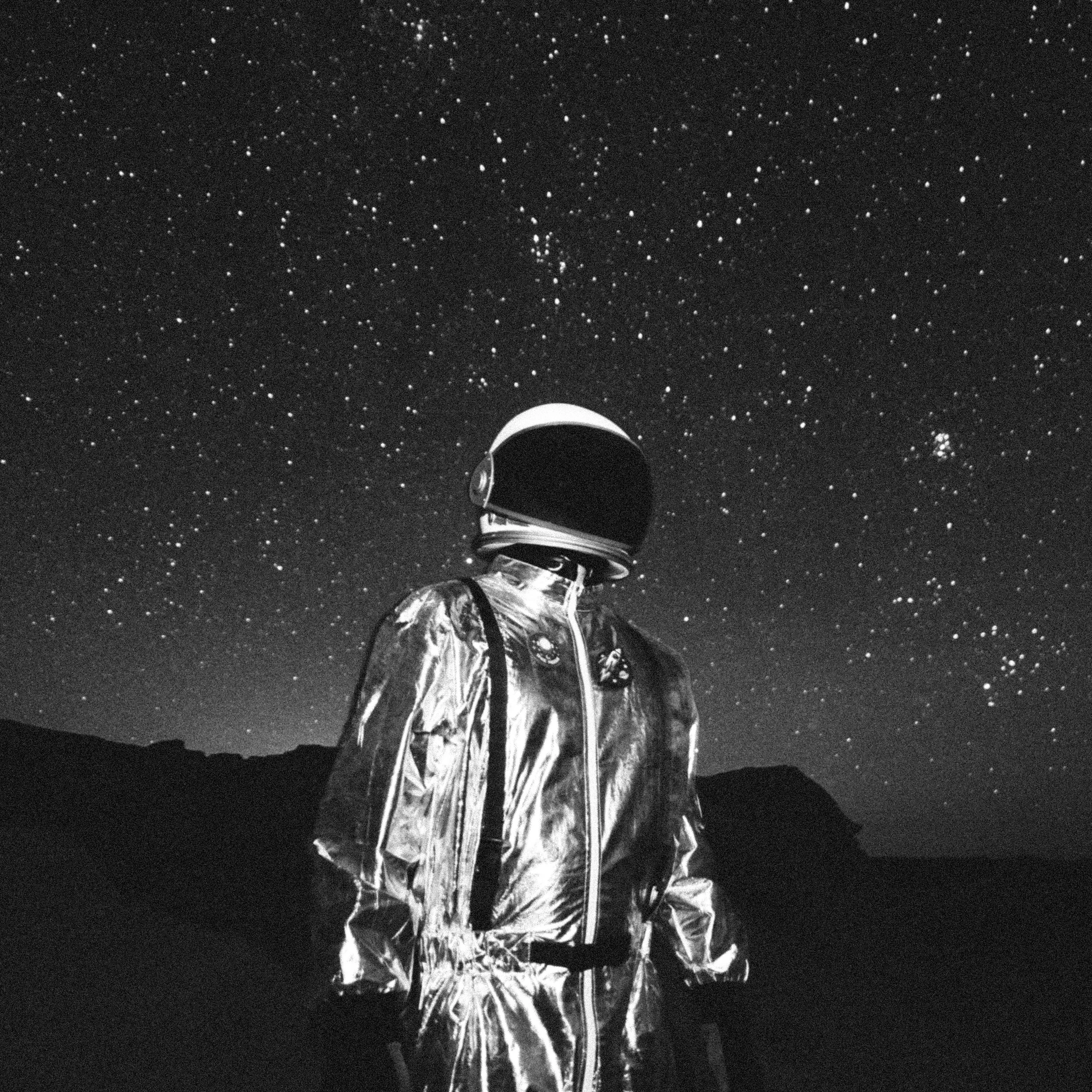 An astronaut in a reflective suit and helmet stands outdoors at night, with a starry sky and mountain silhouettes in the background.