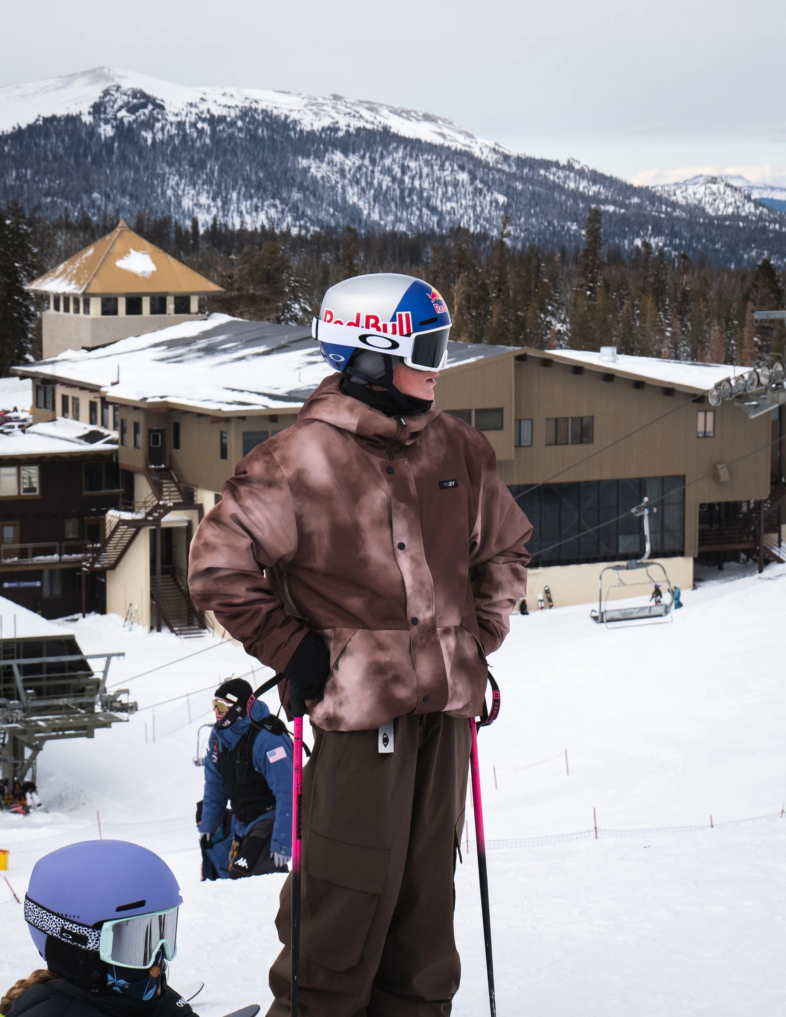 A person wearing a brown jacket, brown pants, and ski gear standing on snow at a ski resort with mountains and buildings in the background.