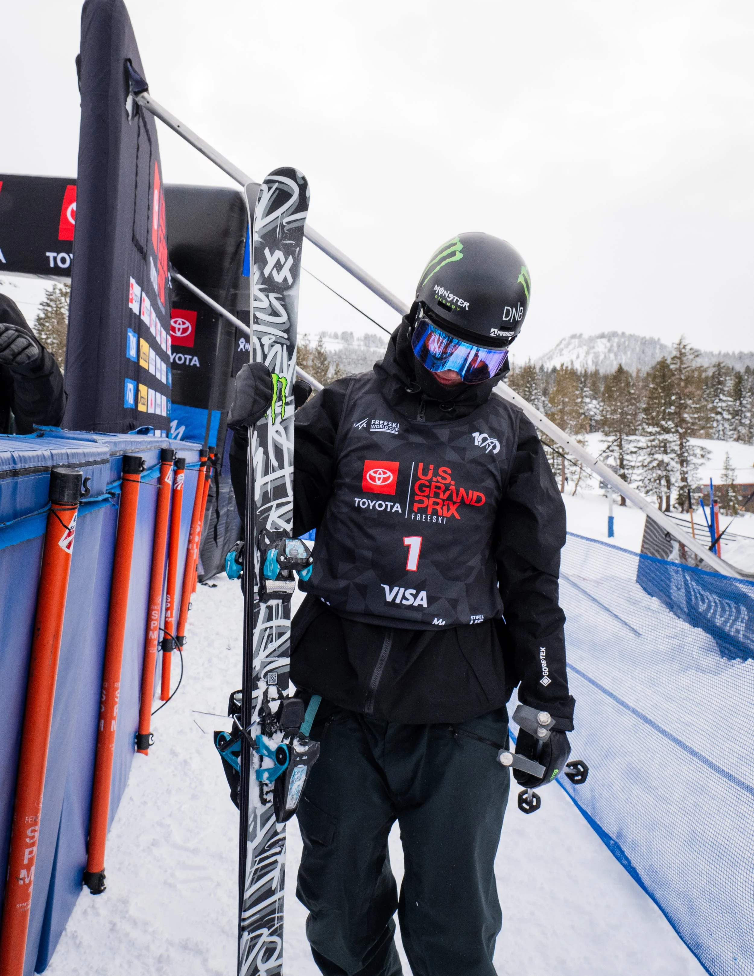 A skier wearing a black helmet with green accents, goggles, and a black jacket with logos stands with skis on a snowy course in a mountain setting, holding ski poles.
