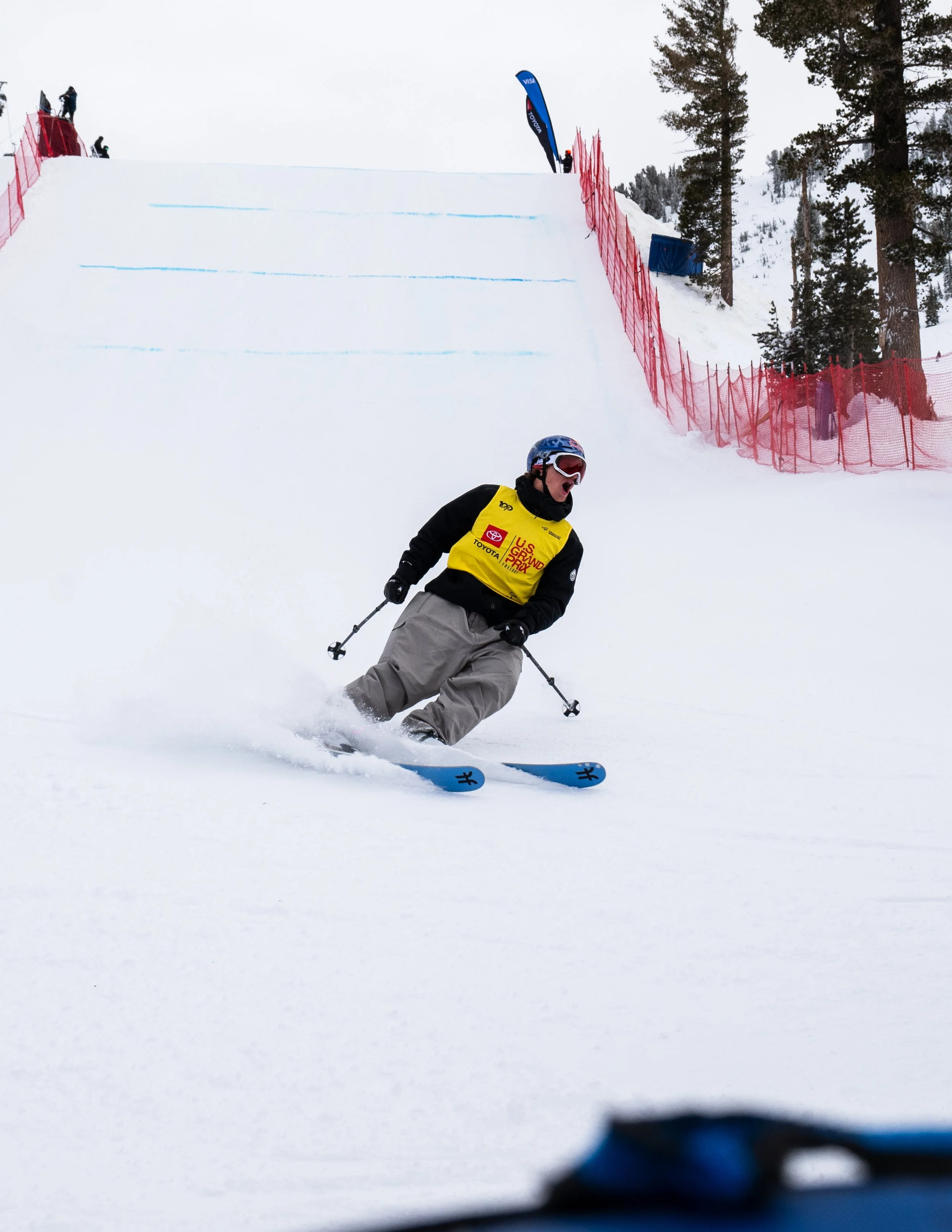 A skier in a yellow bib speeds down a snowy slope during a ski competition or event, with a ski jump and red safety fencing in the background.
