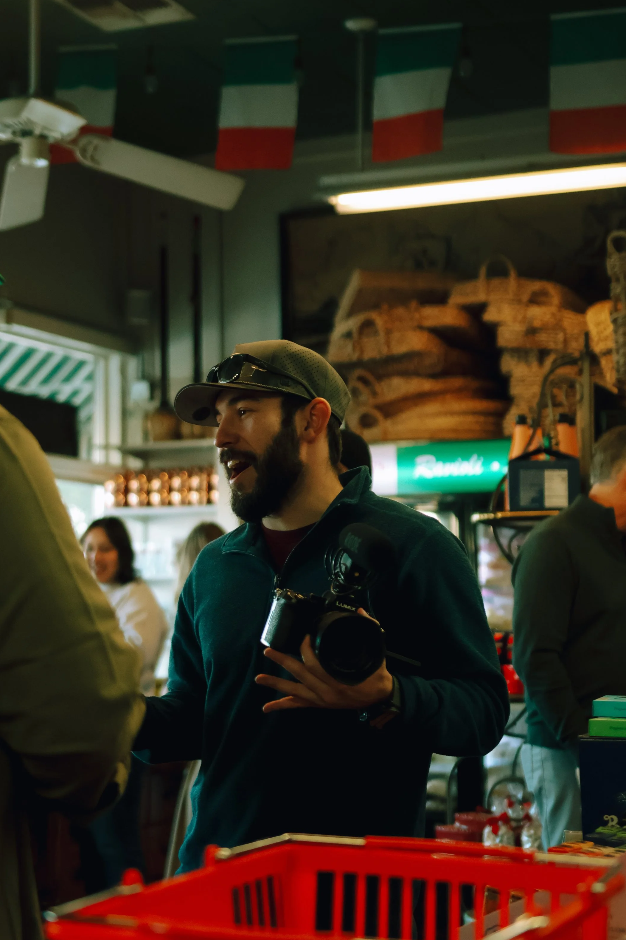 A man with a camera, wearing a cap and sunglasses, talking to someone in a crowded indoor store or cafe. There are people and various items in the background.