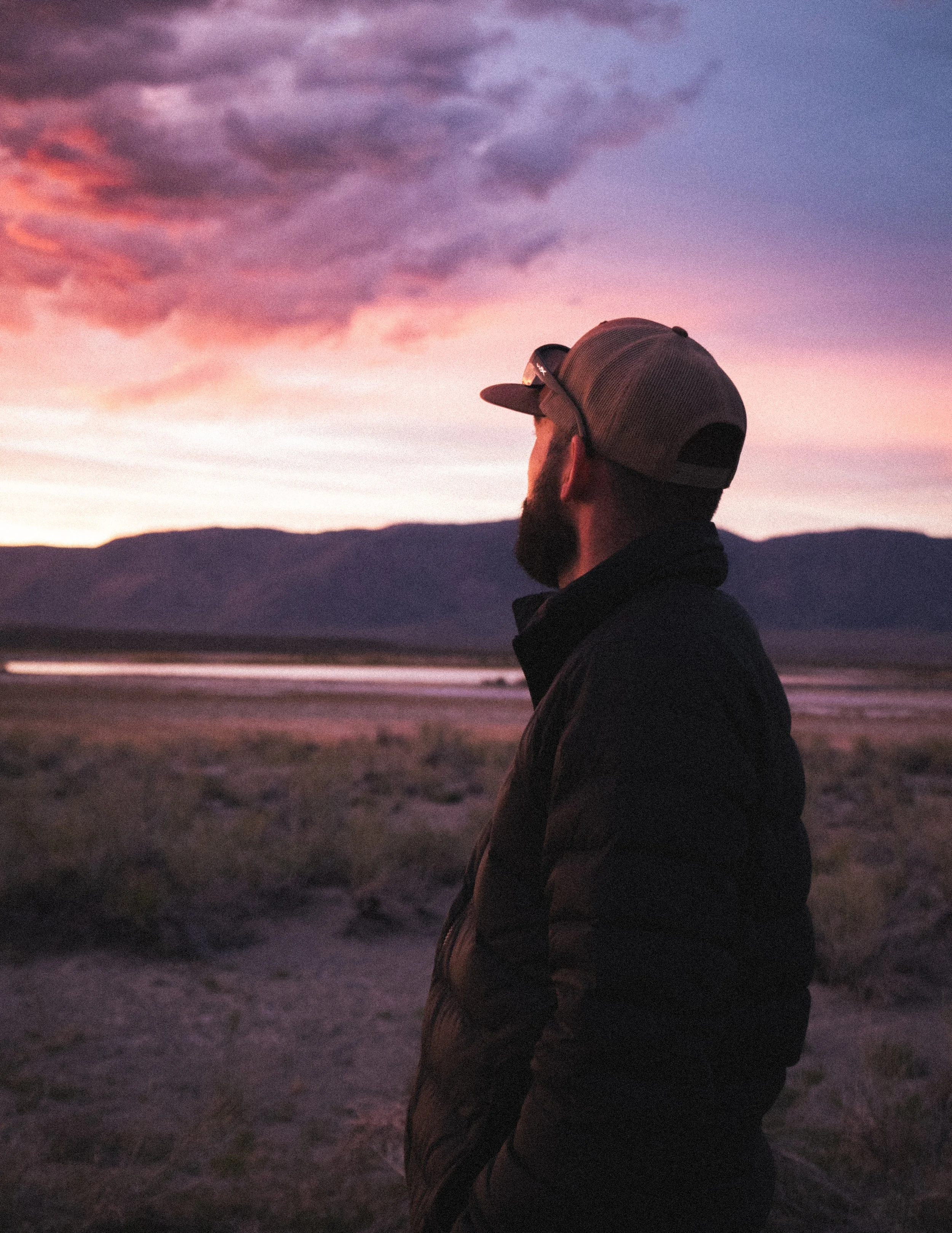 A man (Andrew Sacchini) with a beard wearing a baseball cap and puffy jacket stands outdoors, watching a sunset over mountains and a body of water.