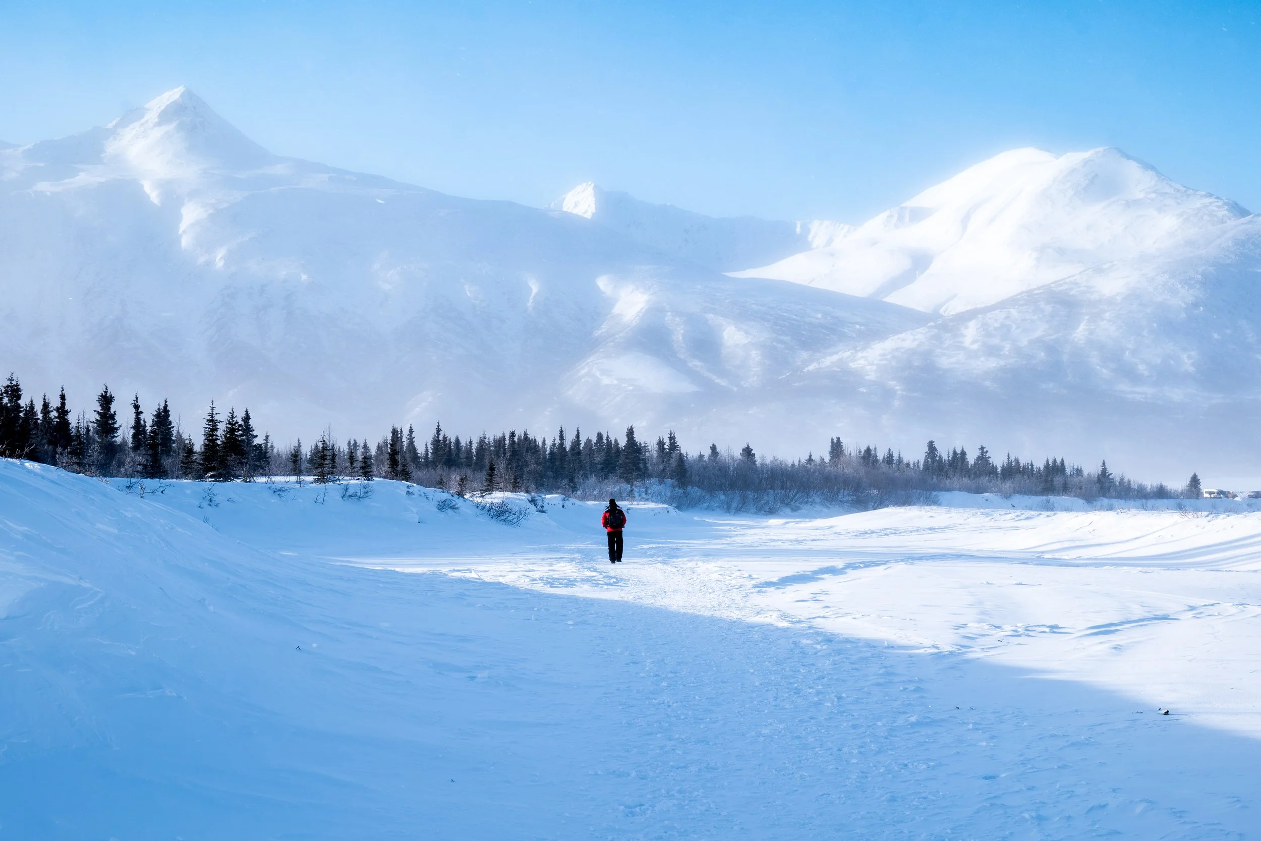 A person walking in a snow-covered landscape with mountains in the background and a clear blue sky.