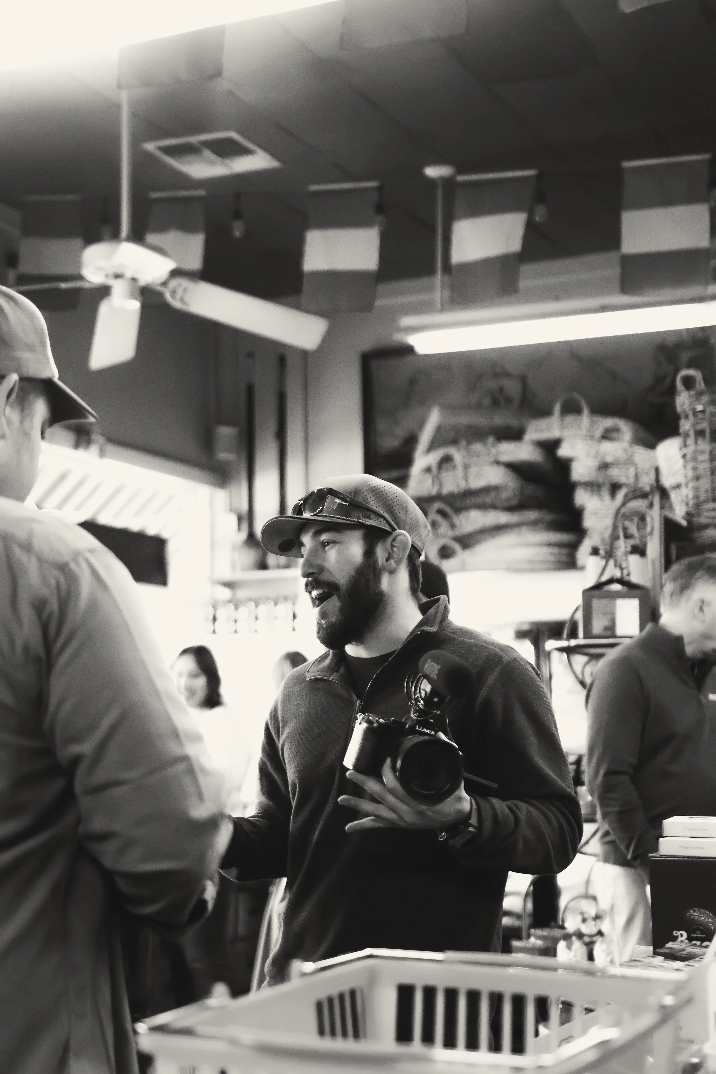 Man with a beard (Andrew Sacchini) wearing a cap and sunglasses on his head holding a camera, talking to another person in a grocery store.