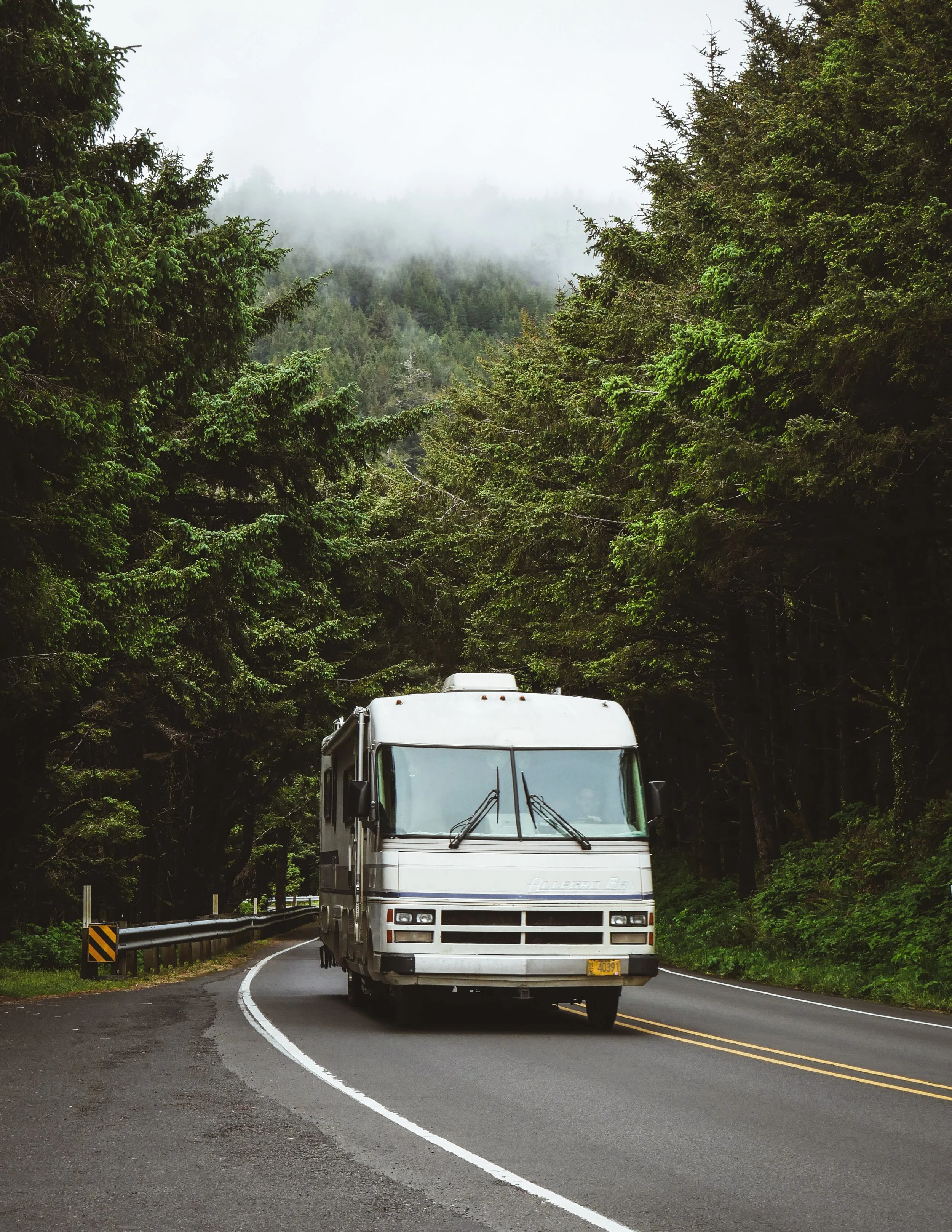 A recreational vehicle driving on a winding forest road surrounded by tall evergreen trees and misty mountains in the background.