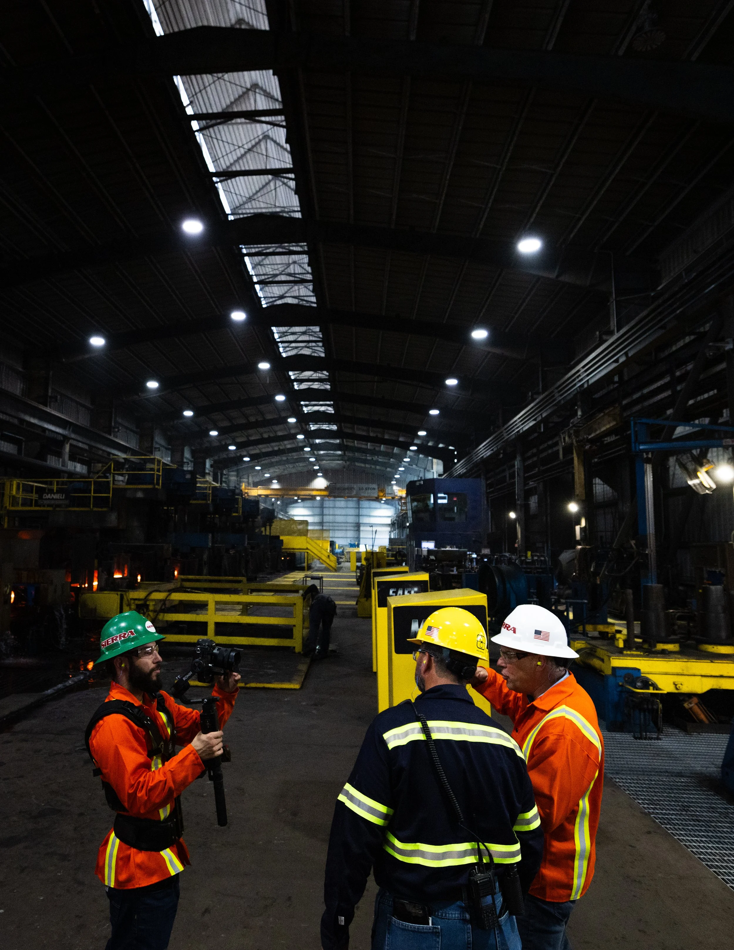 Three industrial workers in safety gear, including helmets and reflective clothing, having a discussion inside a large manufacturing or processing plant.