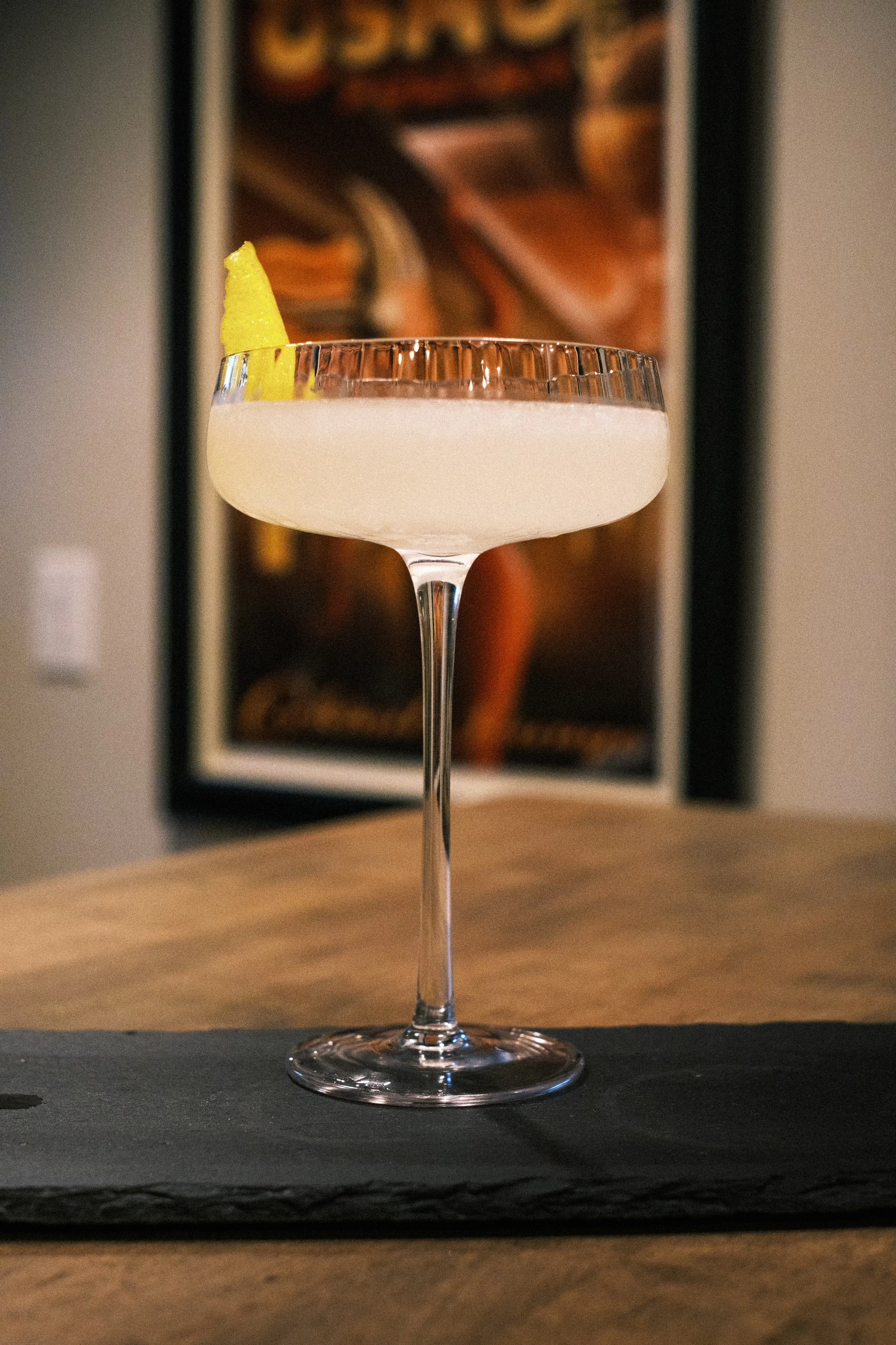 Cocktail in a coupe glass with a lemon twist garnish on a table.