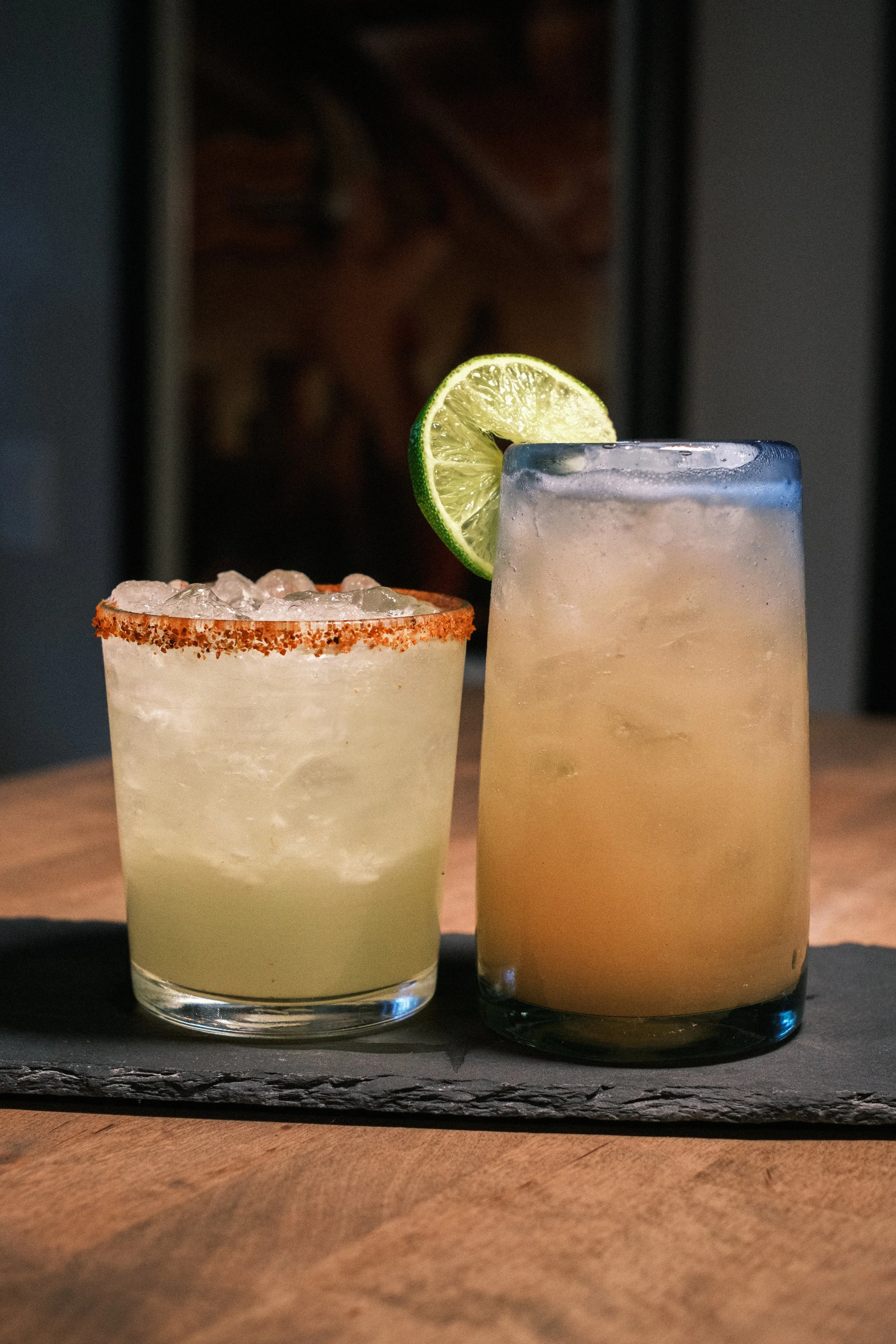 Two cocktails on a slate tray; the one on the left has a chili-rimmed glass, the one on the right is garnished with a lime slice.