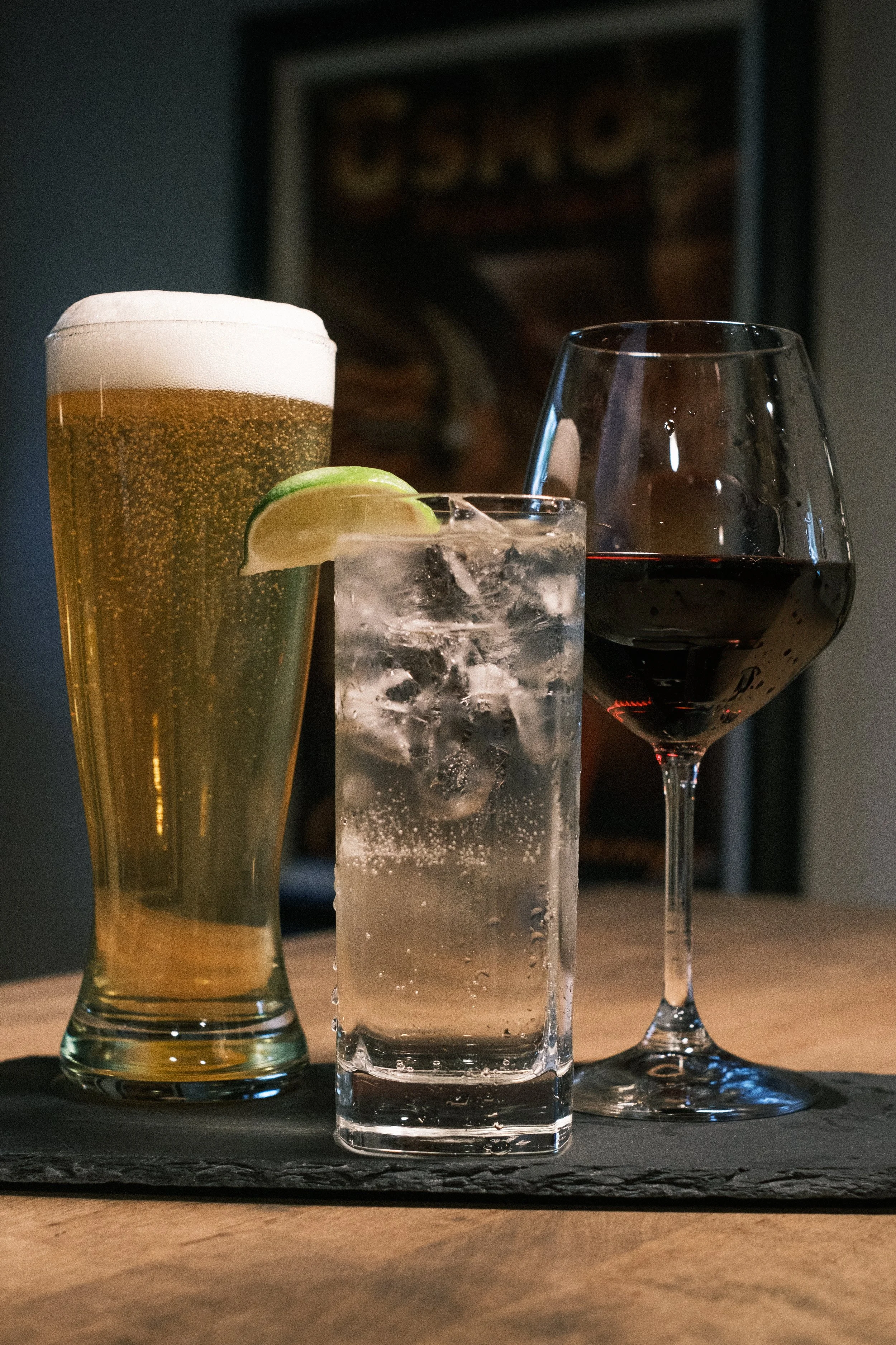 Three drinks on a table: a pint of beer with foam, a glass of soda with lime, and a glass of red wine on a slate serving tray.