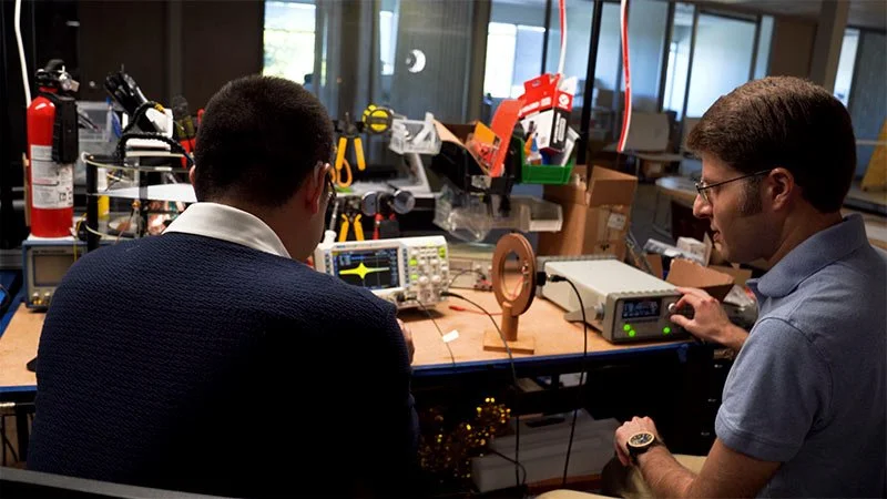 The founders of Etherdyne working at a cluttered electronics workbench with experimental equipment and tools, including oscilloscopes, a small fan, and a fire extinguisher, in the Etherdyne Lab.