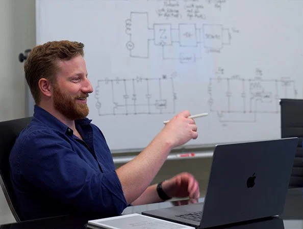 A man with a beard in a blue shirt sitting at a desk with a laptop, pointing at a whiteboard with electrical circuit diagrams written on it.